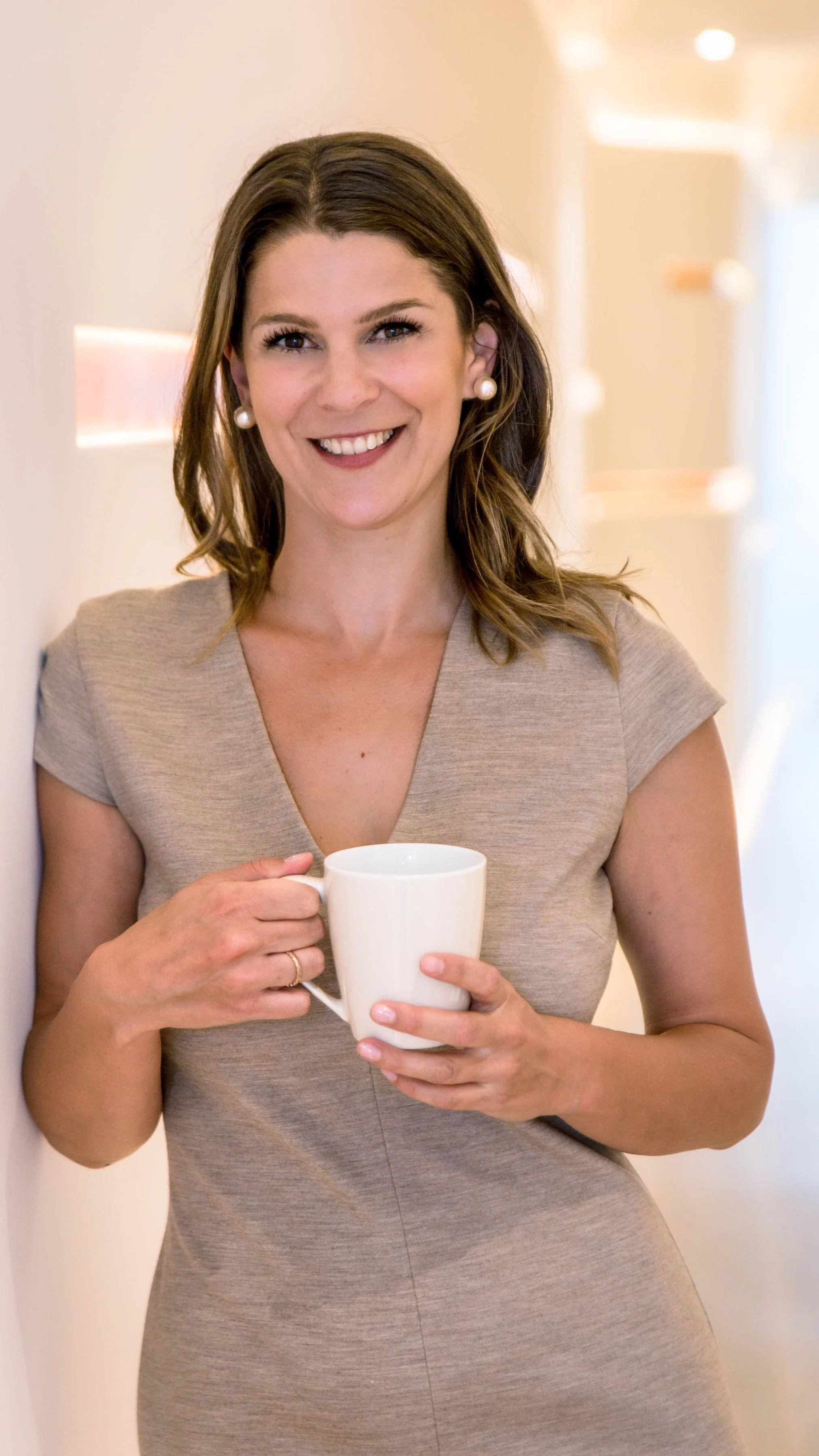 Eine Frau lehnt an einer Wand und hält eine Tasse. Sie lächelt und trägt ein beigefarbenes Kleid.