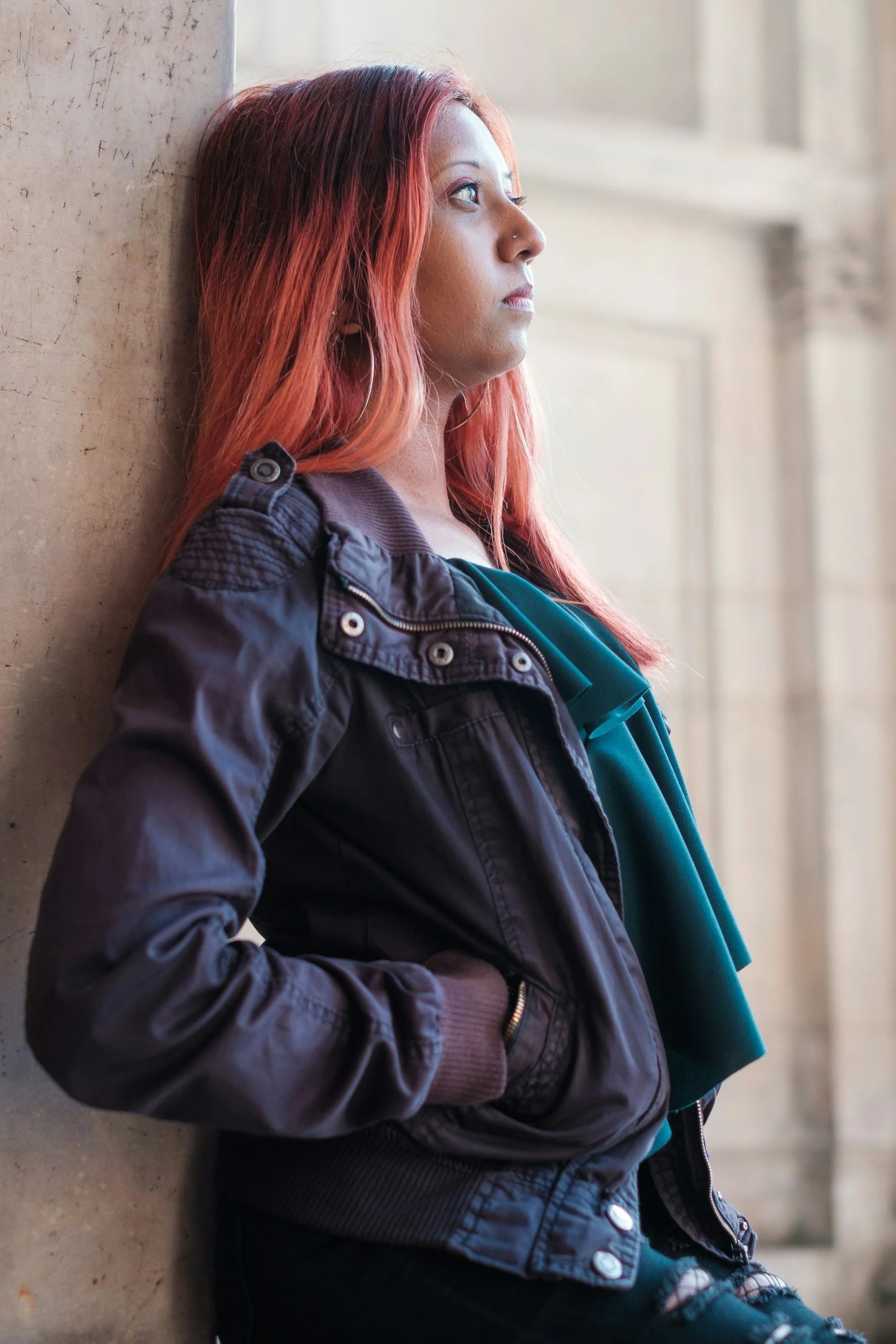 South Asian Person with long red hair leaning against a wall, wearing a dark jacket and green top.