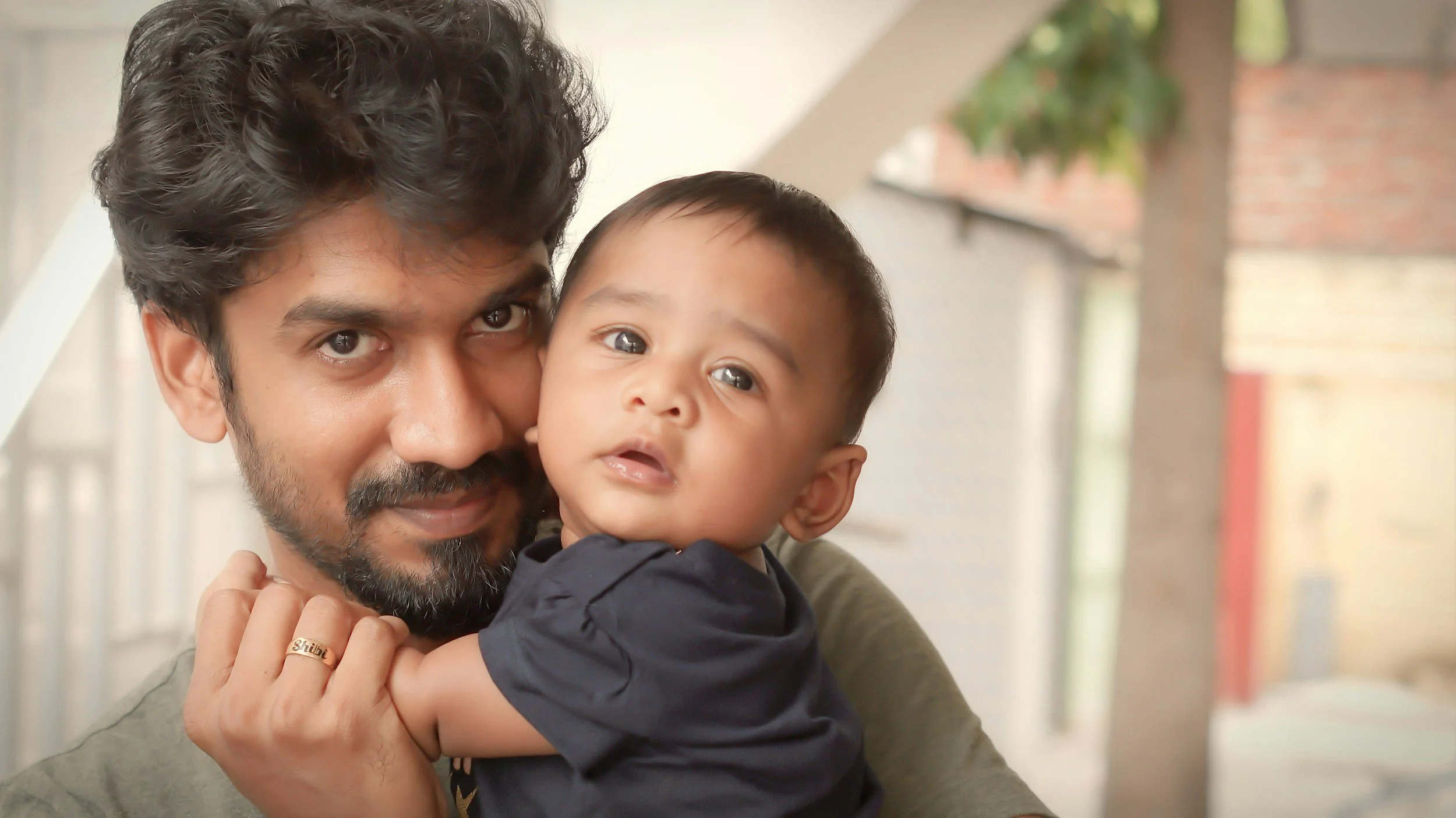 Indian Man holding a baby indoors, both looking at the camera.