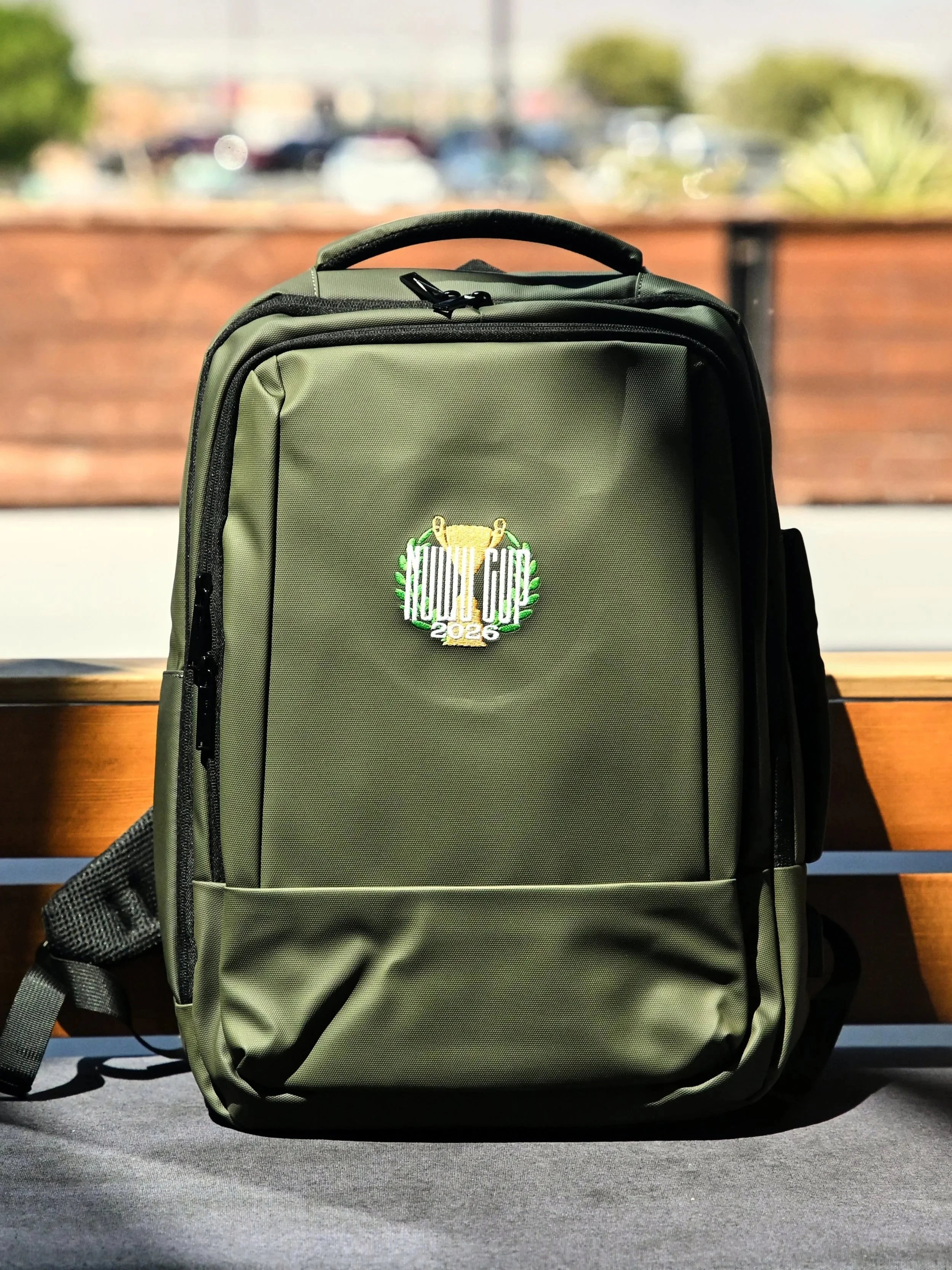 An olive green NuWu Cup 2026 official judge backpack with a custom embroidered trophy patch, sitting outdoors on a wooden bench at NuWu North.
