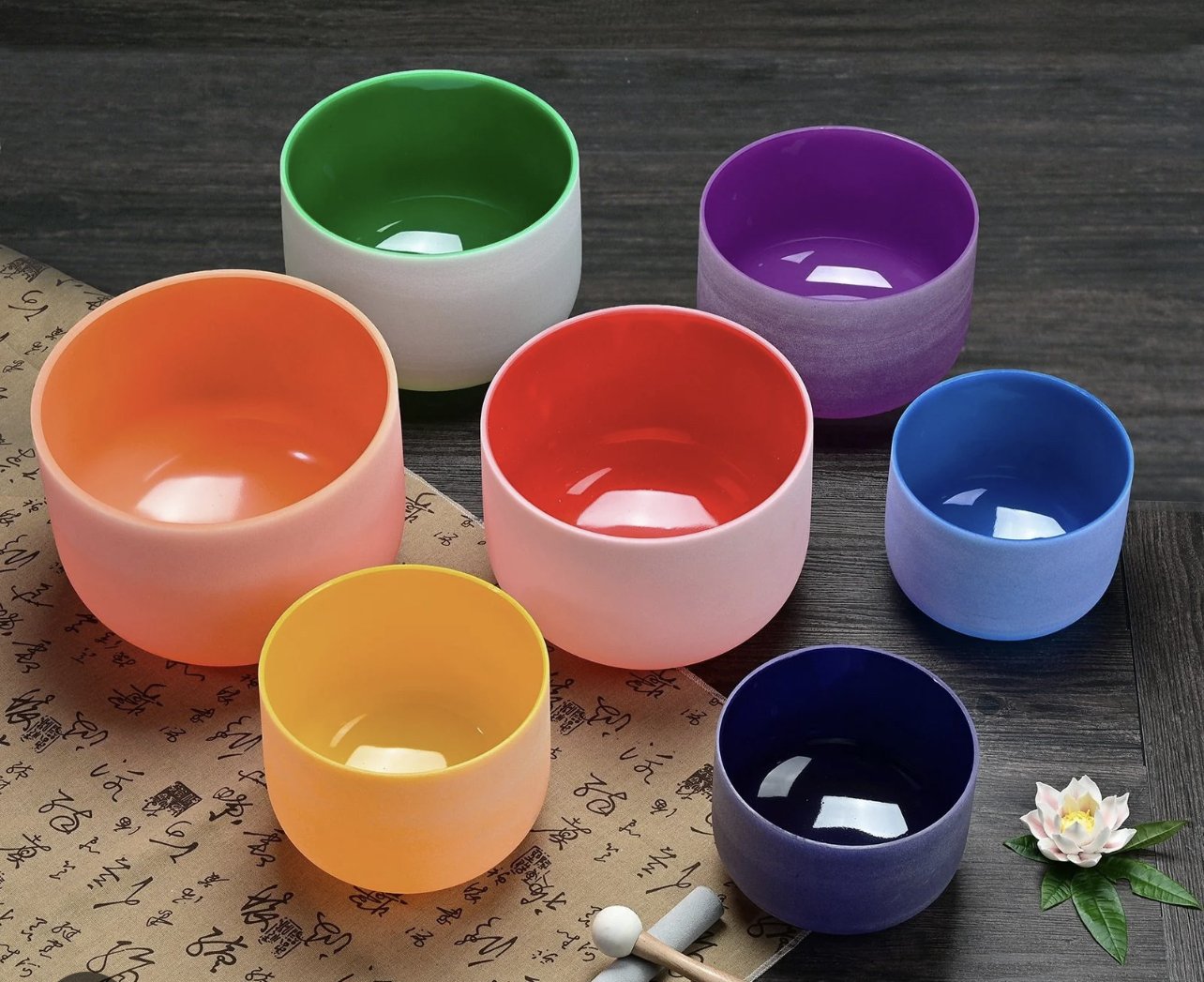 Colorful glass bowls in green, purple, orange, red, yellow, blue, and dark purple arranged on a dark wood surface, with a rolled scroll and a flower with green leaves nearby.