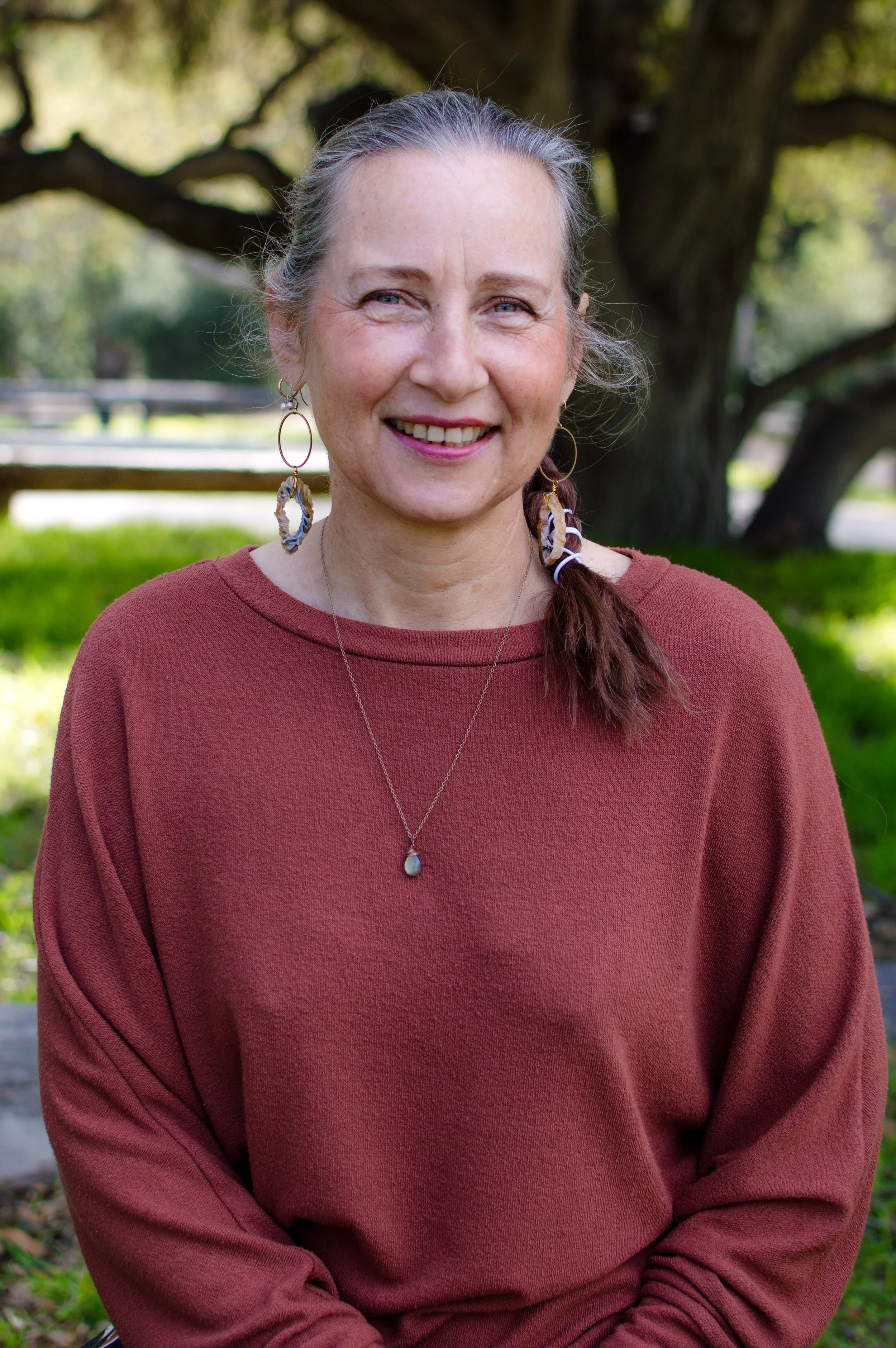 A smiling woman with gray hair in a low ponytail, wearing a rust-colored long-sleeve shirt, earrings, and a necklace, standing outdoors in front of a large tree and green grass.
