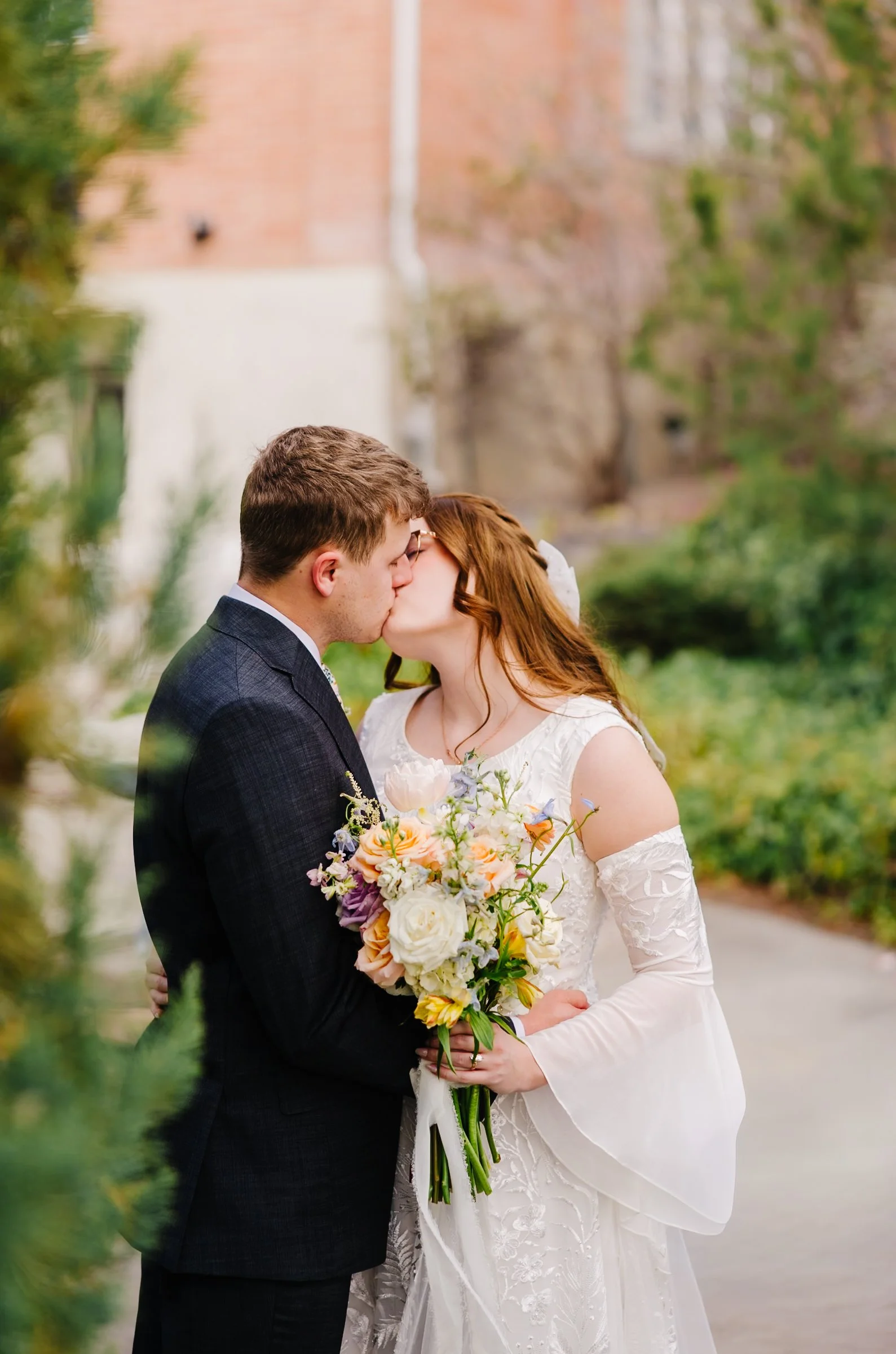 A bride and groom kiss outdoors during their wedding, holding hands, with scenic mountains and cloudy sky in the background.