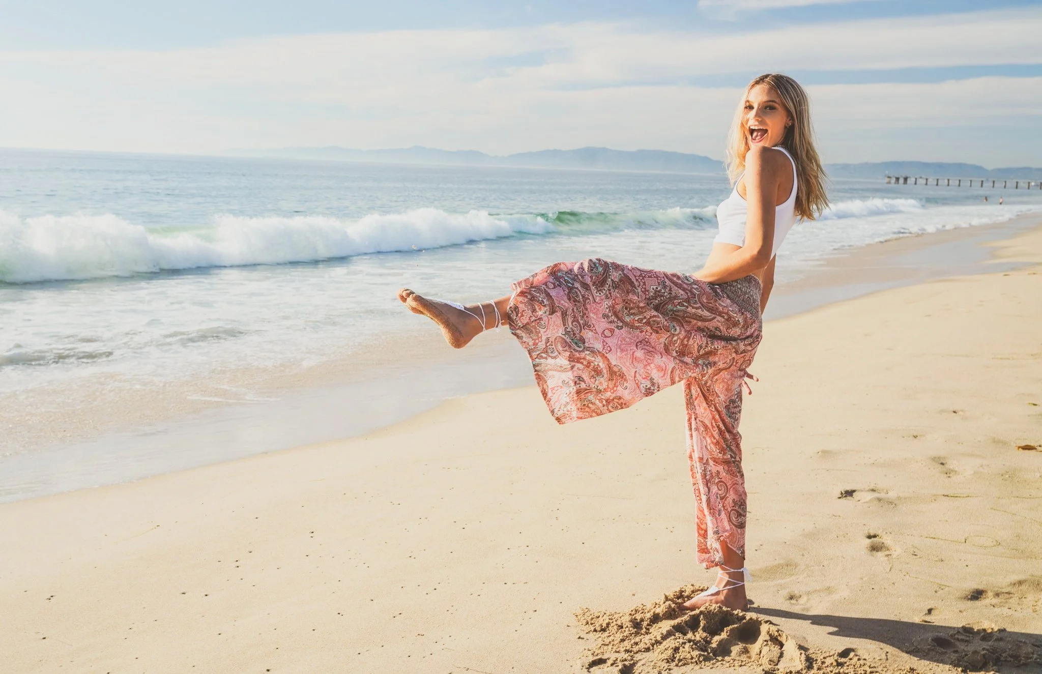 A woman standing on a sandy beach, smiling with one leg raised, wearing a white tank top and a floral patterned skirt, with ocean waves and a pier in the background.