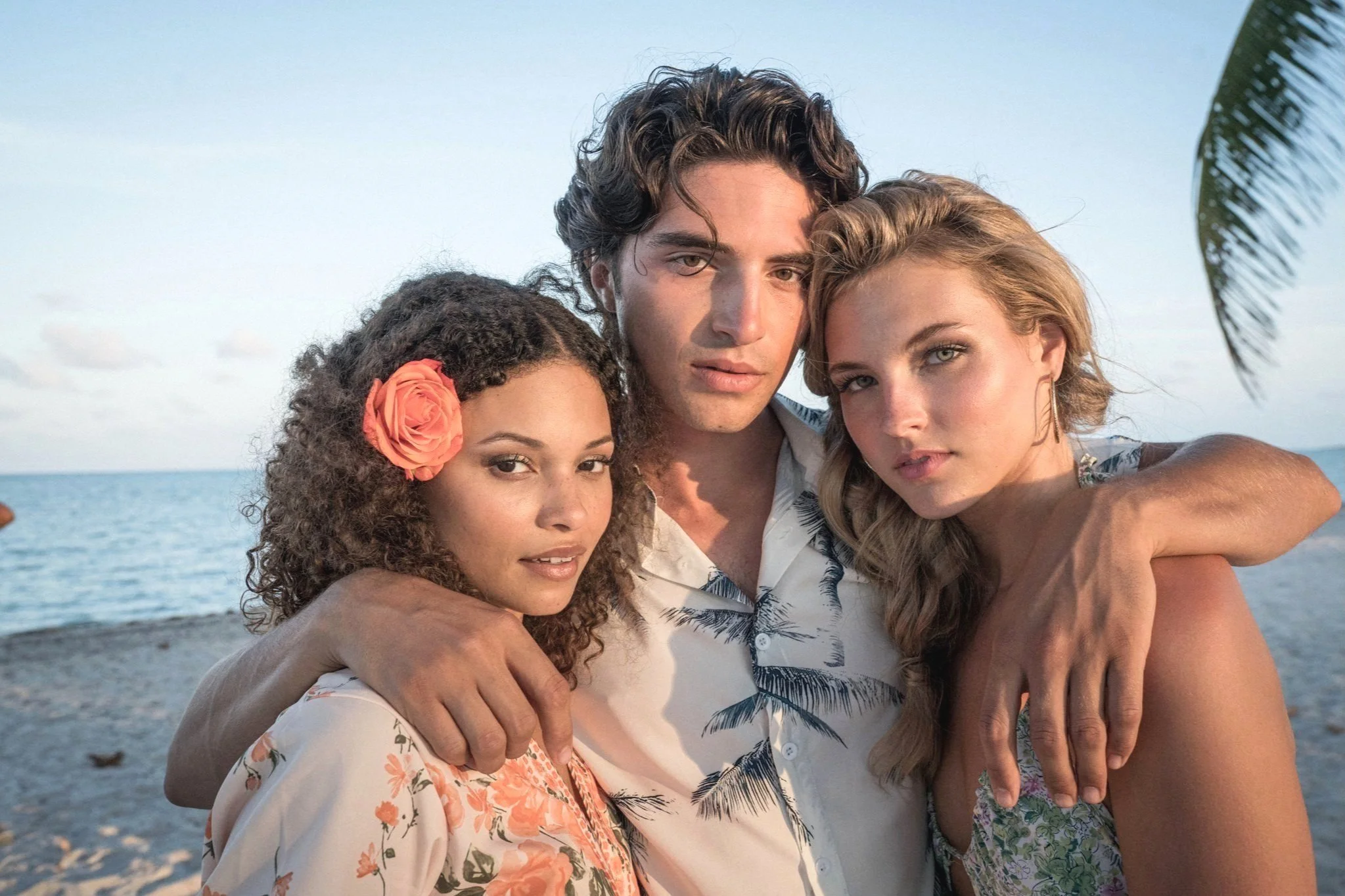 Three young adults, two women and one man, standing close together on a beach with the ocean in the background, embracing each other and looking at the camera.