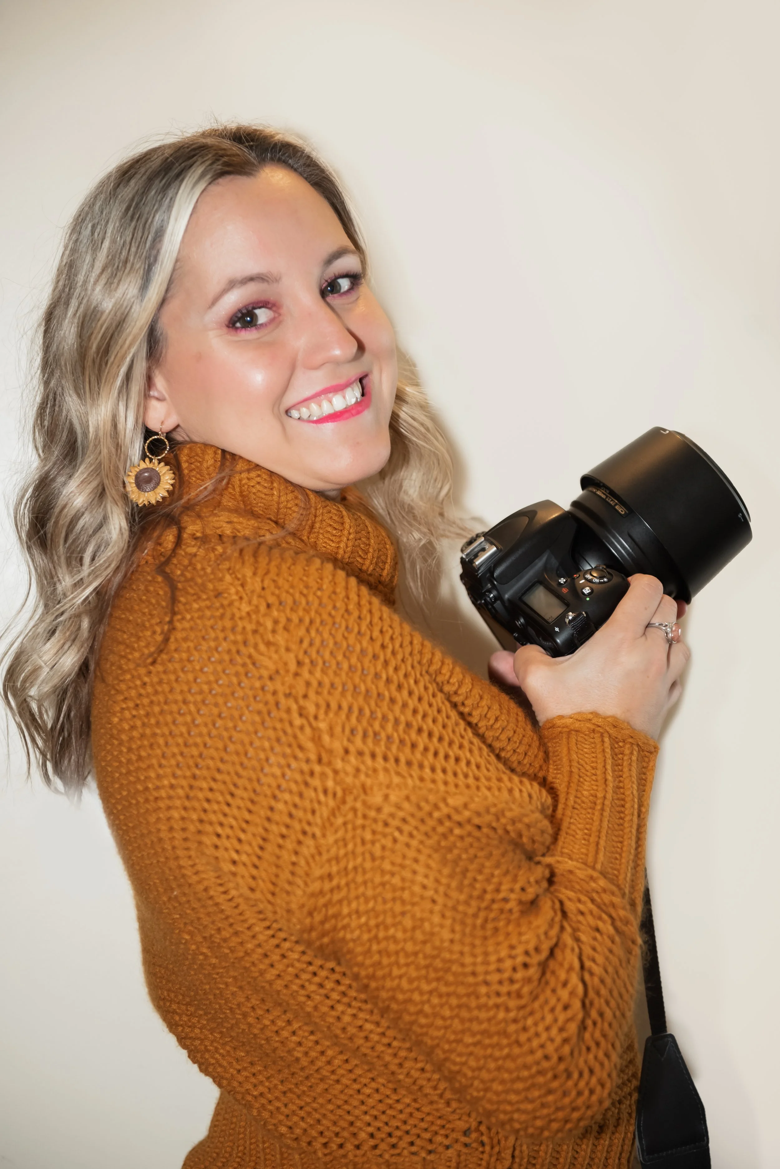 A woman with blonde wavy hair holding a camera and smiling, wearing a rust-colored knit sweater and sunflower-shaped earrings.
