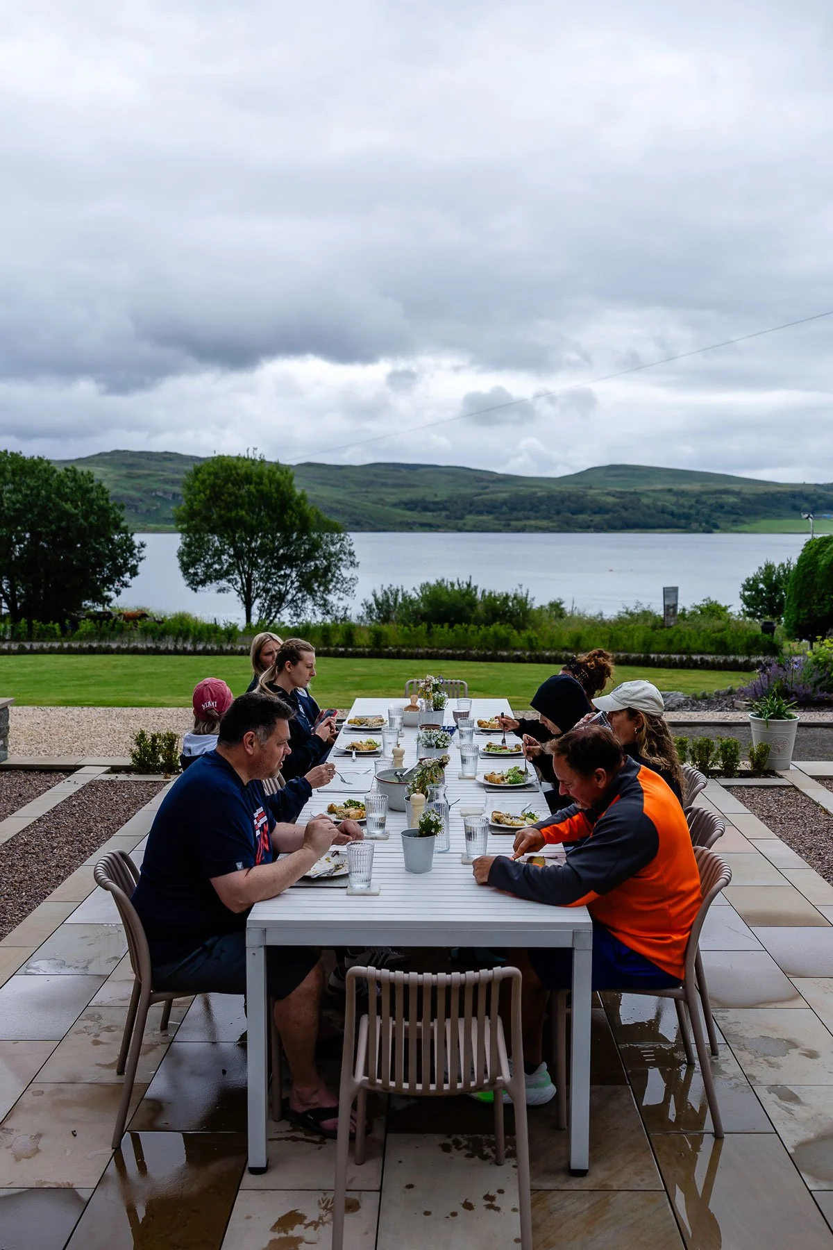 Family and friends gathered around an outdoor dining table enjoying a meal with a scenic lake and green hills in the background on a cloudy day.