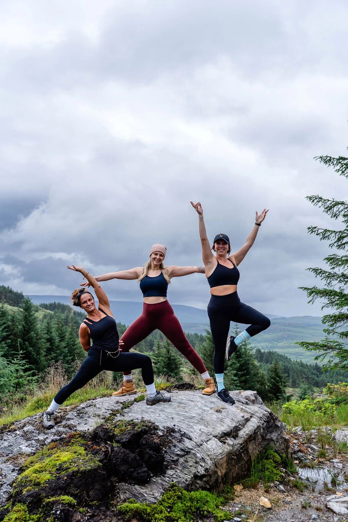 Three women in athletic clothing posing on a large rock in a mountain landscape with trees and cloudy sky.