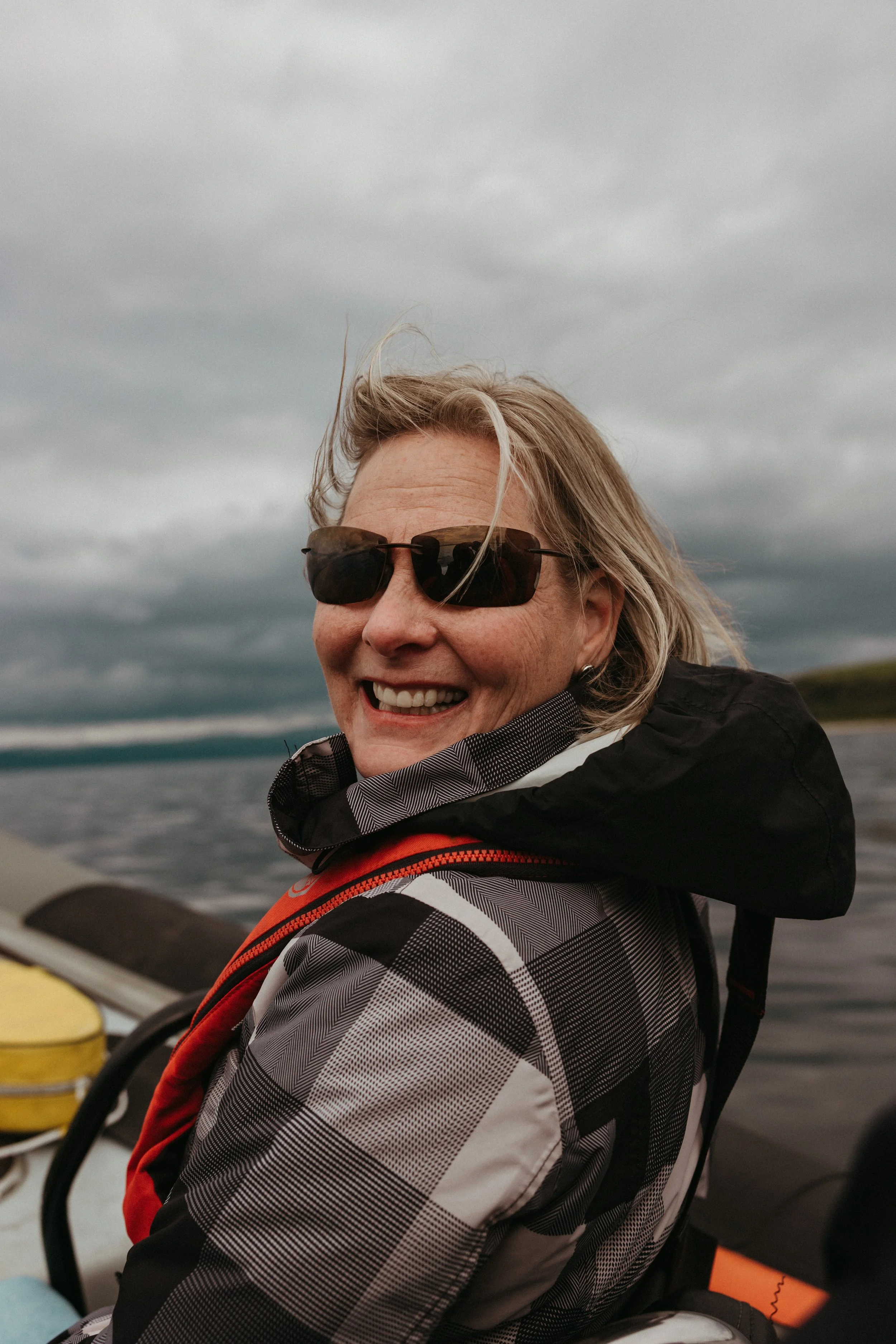 A smiling woman wearing sunglasses, a checkered jacket, and a life jacket, on a boat on a cloudy day with water and distant shoreline in the background.