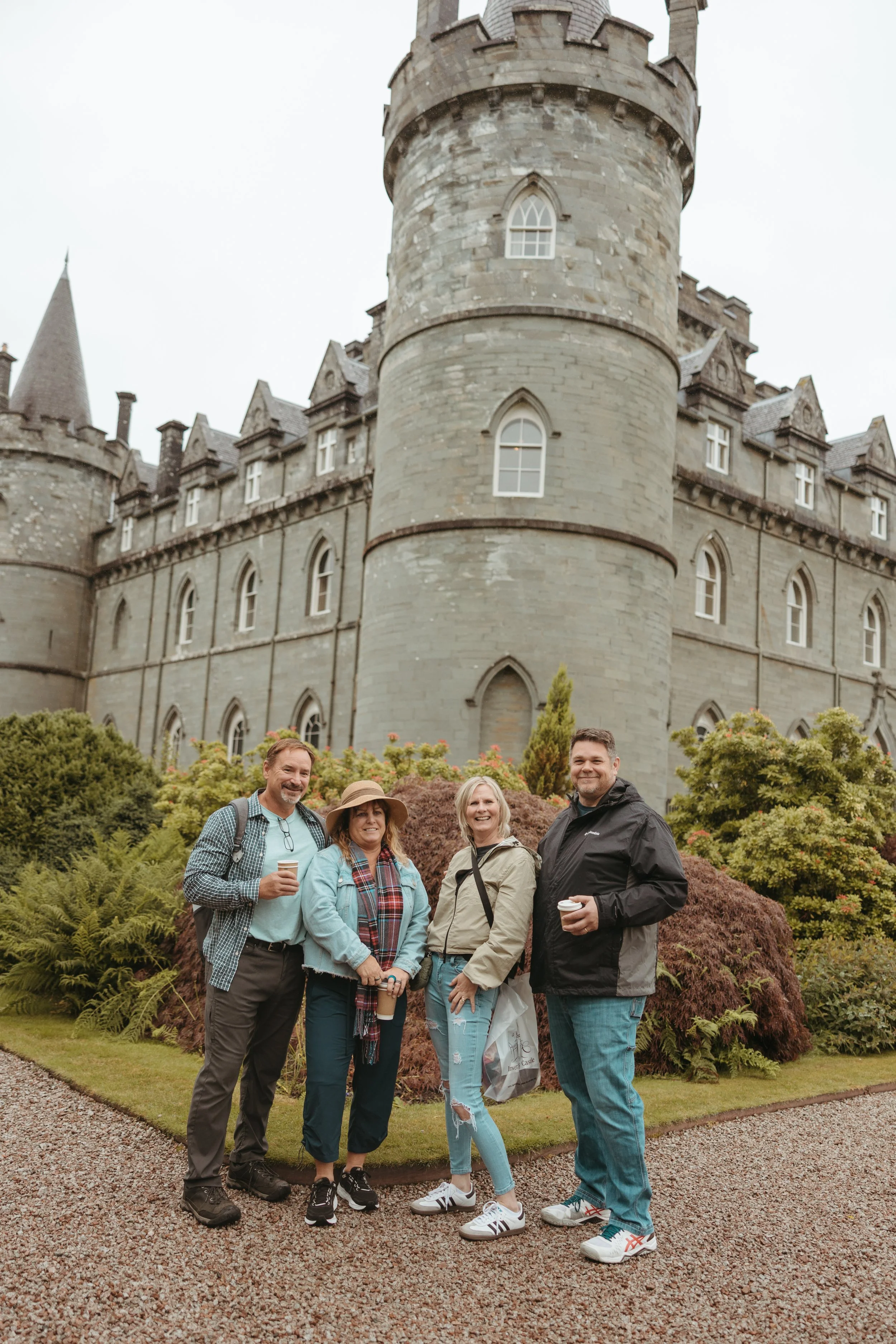 Four people standing in front of a castle, smiling, with greenery around them, and holding coffee cups.