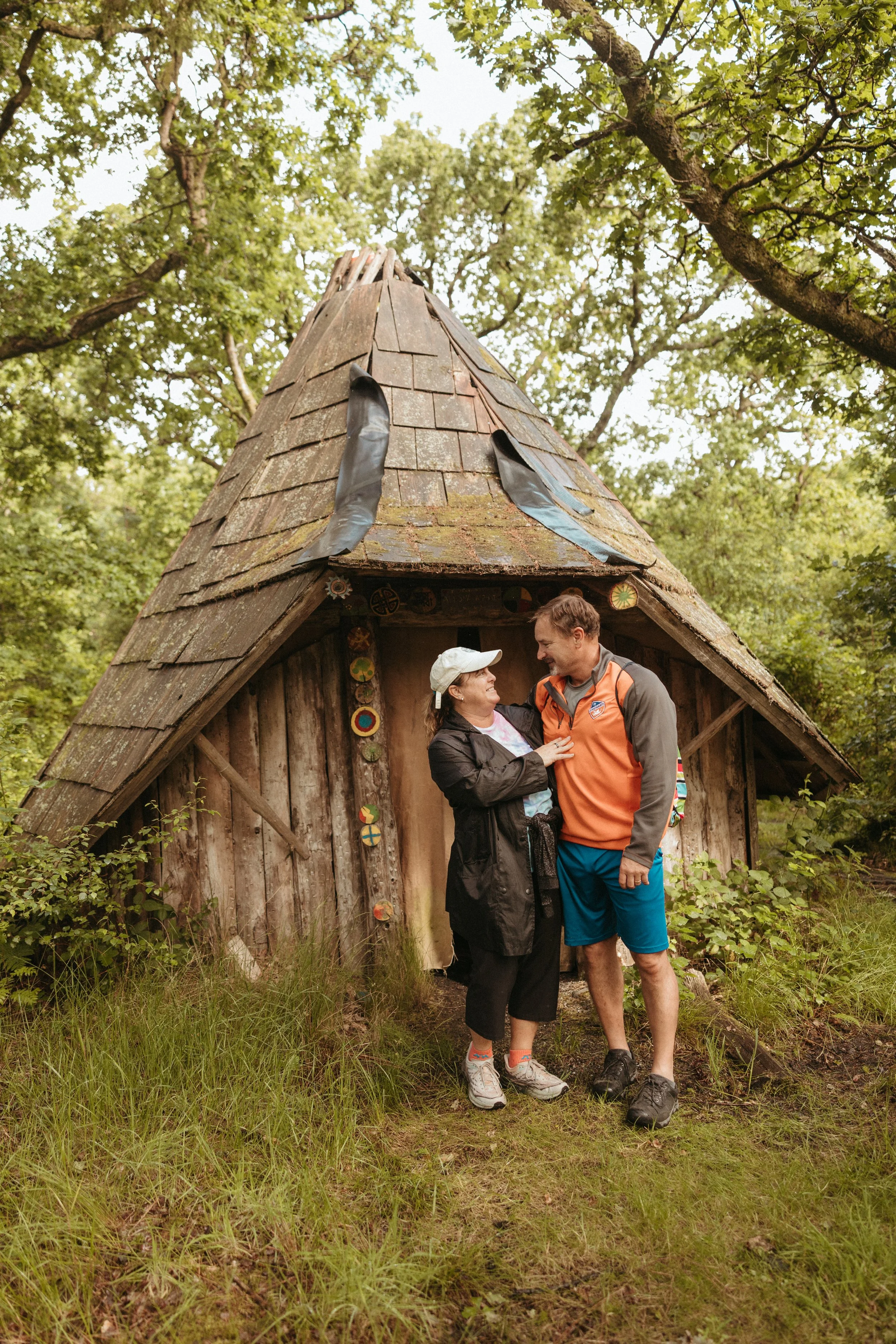 A man and woman smiling together outside a small, rustic wooden hut in a wooded area, with the woman wearing a white cap and the man in an orange and grey jacket.