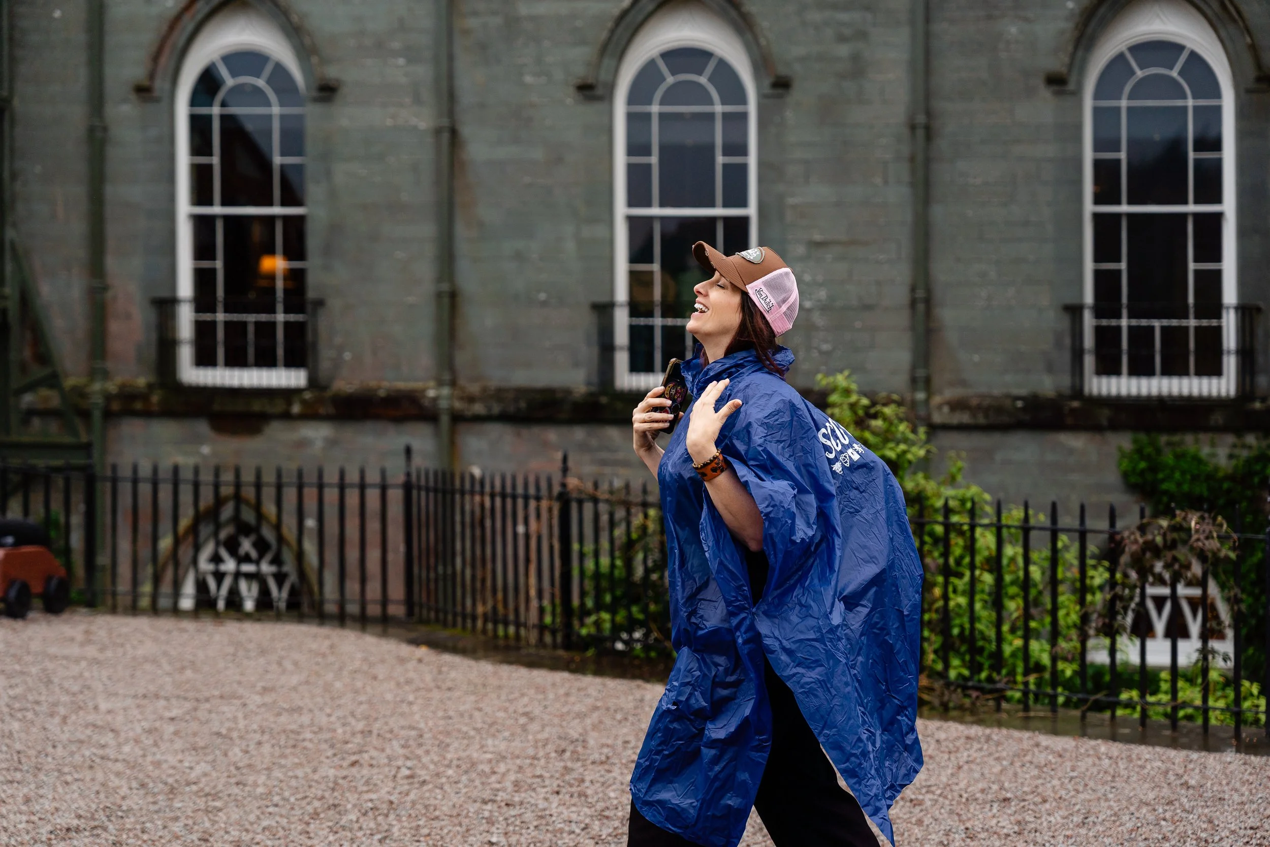 A woman smiling and looking up, wearing a blue rain poncho, a brown cap with patches, and a wristwatch, standing outside in front of a gray building with tall arched windows.
