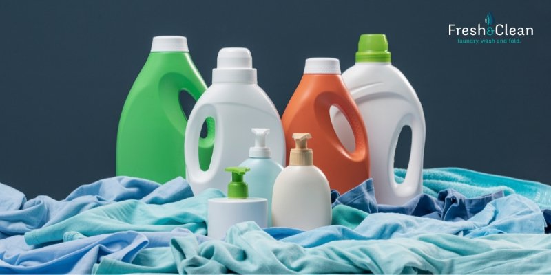 Assortment of laundry detergents and fabric softeners on a surface with folded clothes, with a logo in the top right corner reading 'Fresh & Clean'