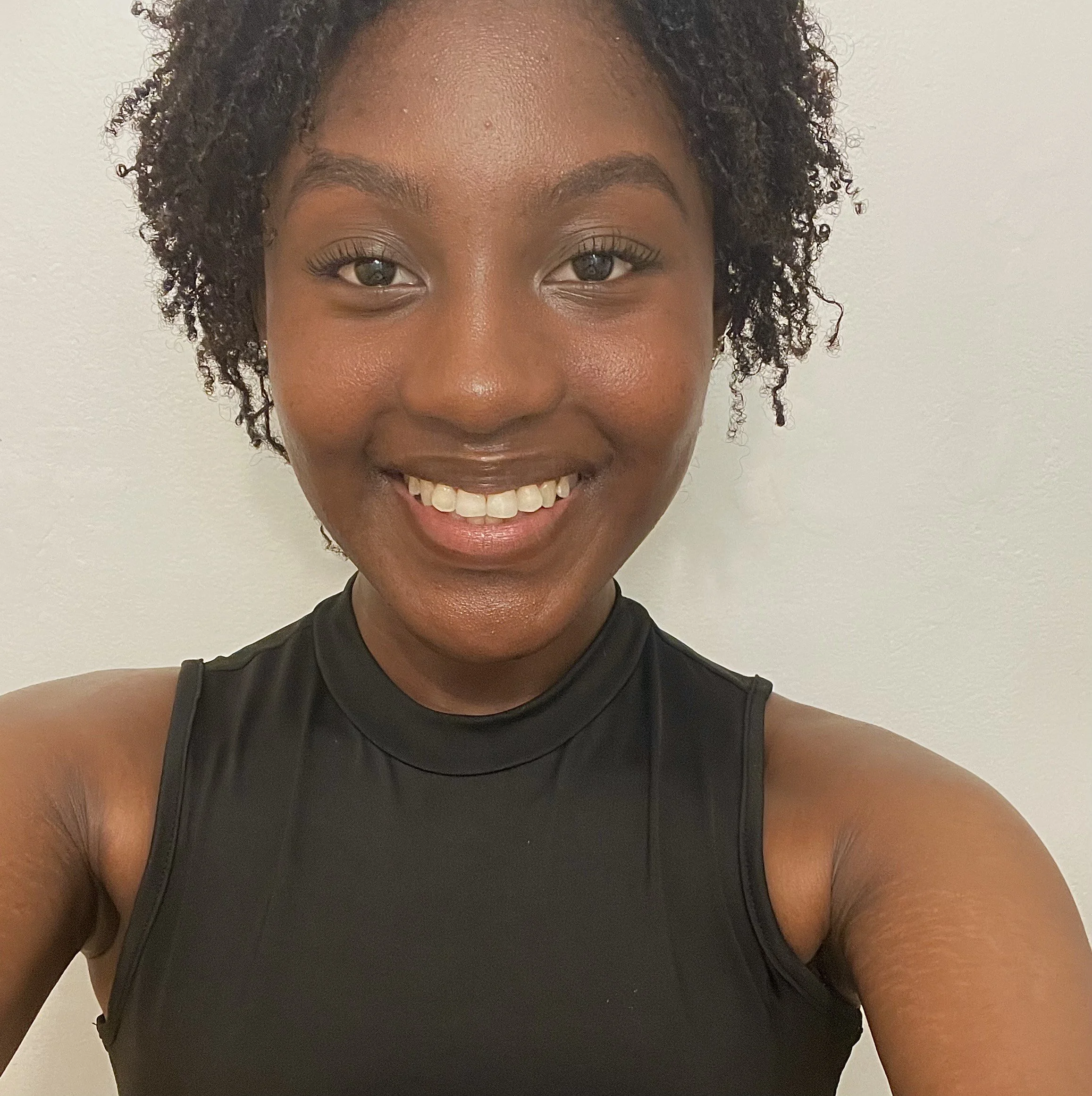 Close-up of a smiling African American woman with short curly hair, wearing a black sleeveless athletic top, against a plain light-colored background.
