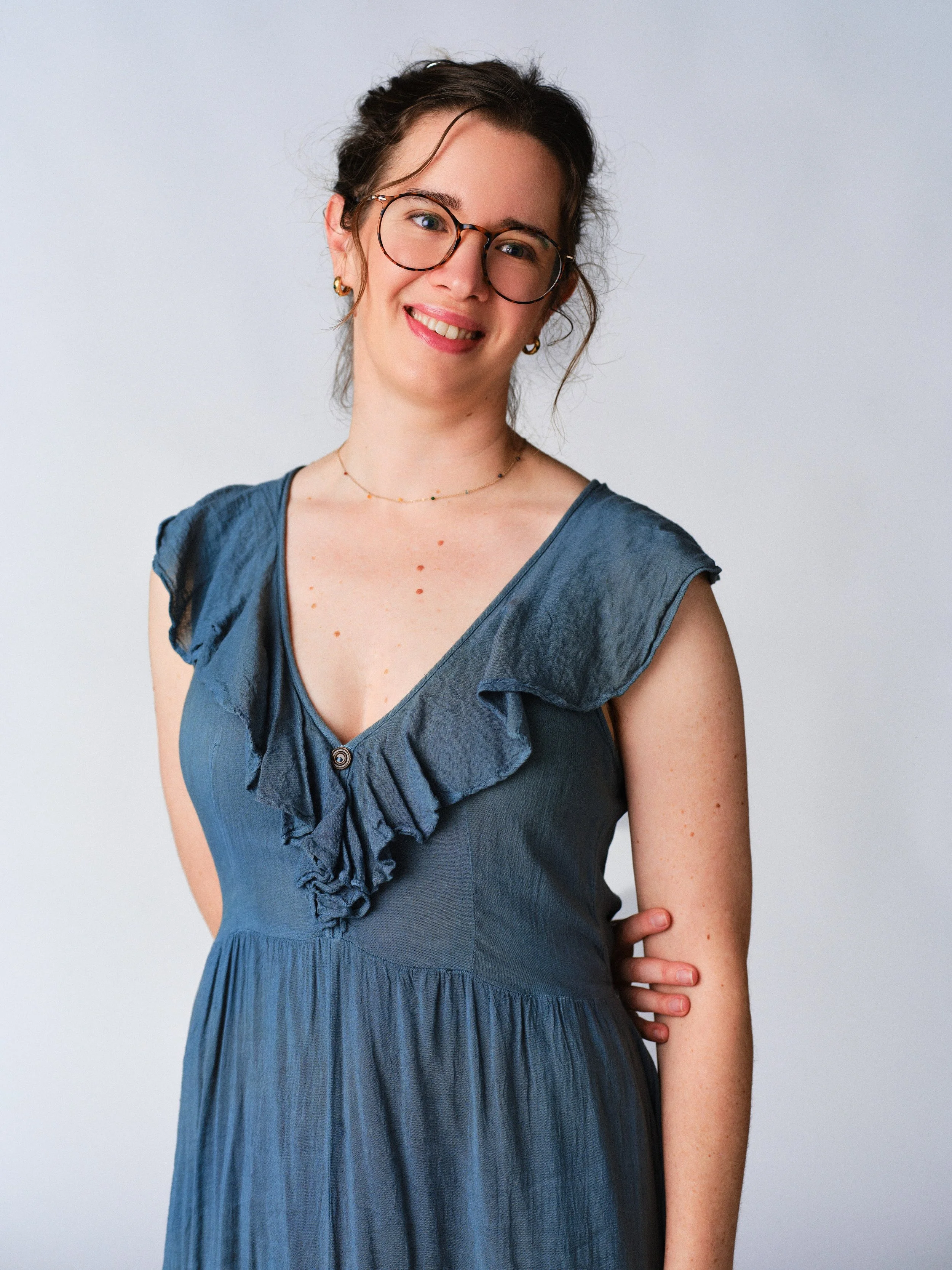 A woman with curly brown hair and glasses standing outdoors by a wooden fence, overlooking a body of water surrounded by greenery and trees.