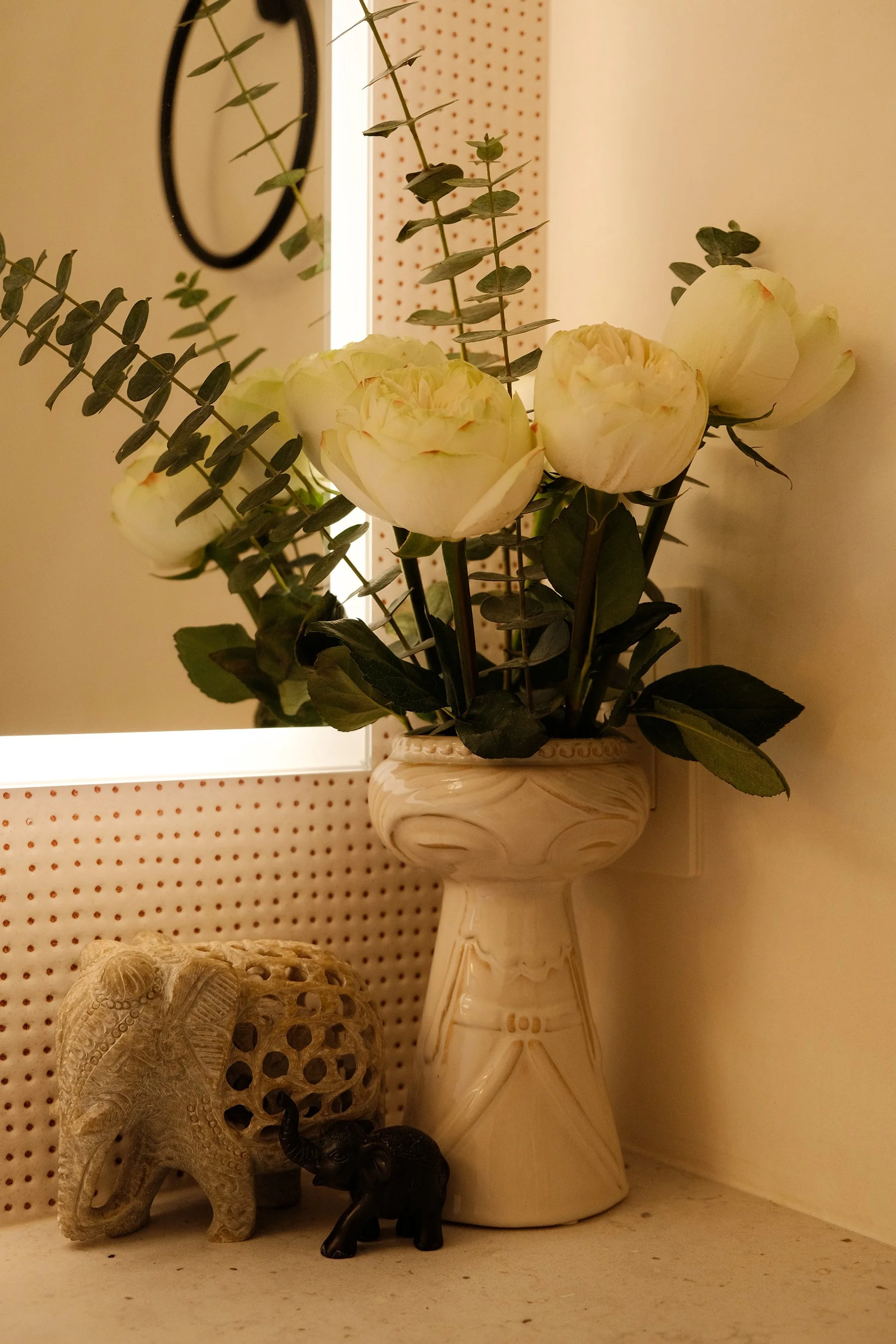 Vase with white peony flowers and greenery on a shelf, with elephant and elephant figurines below, and decorative items in the background.