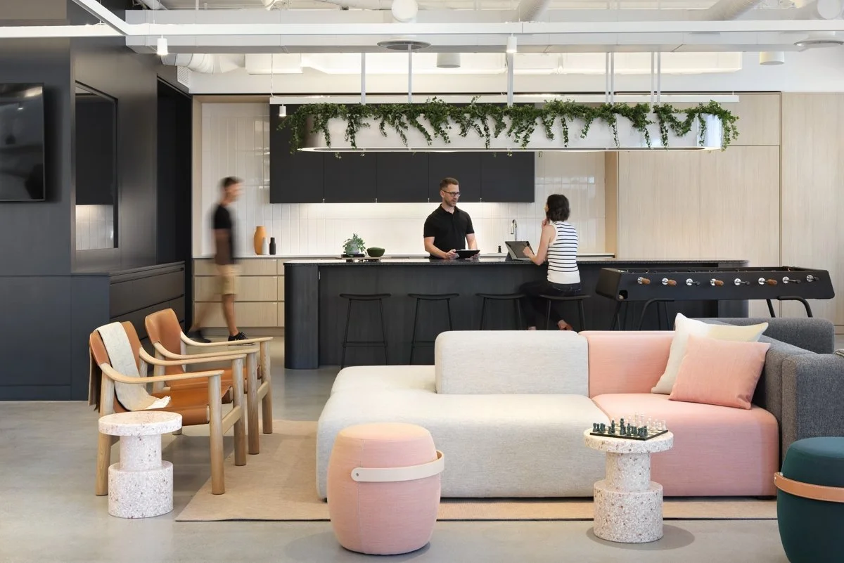 Modern lounge with a kitchen and bar area, seating with pastel and wooden furniture, a foosball table, and decorative green plants hanging above the bar.