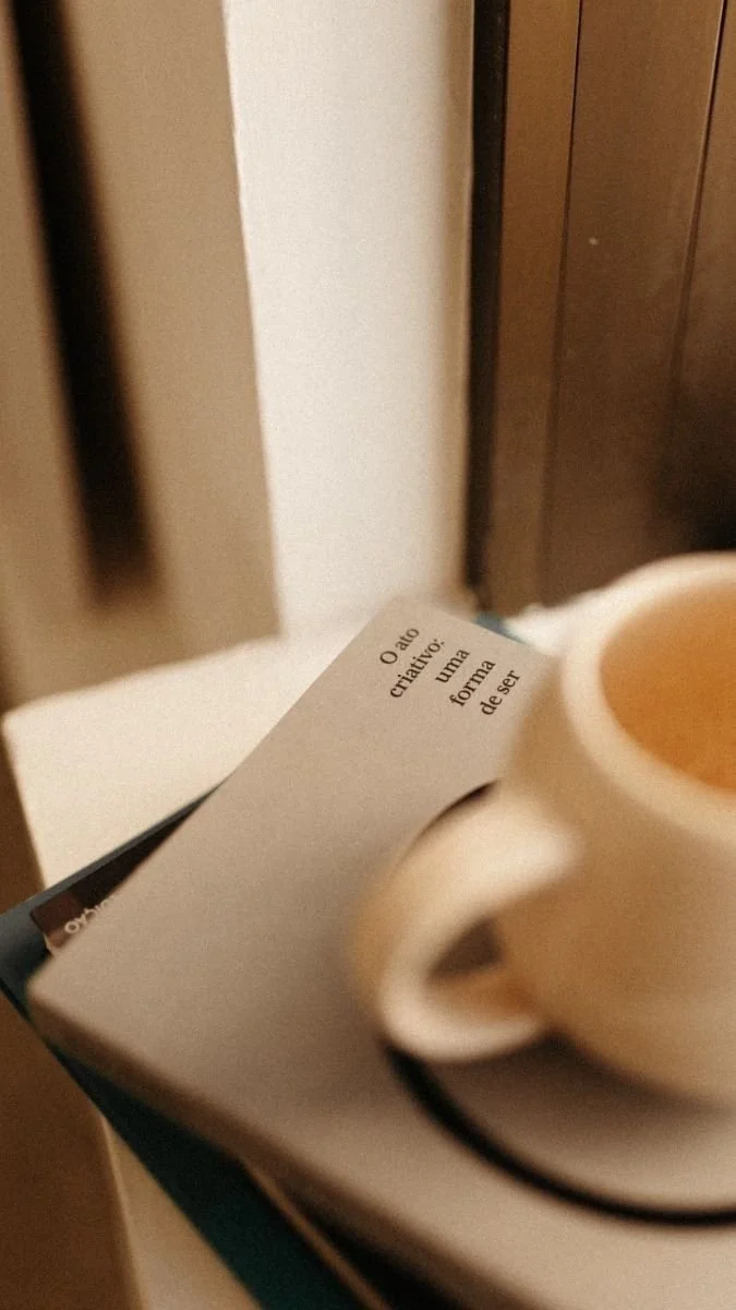 A white coffee cup on a saucer placed on a closed book or notebook, with a partial view of a paper with a Portuguese quote about creative acts.