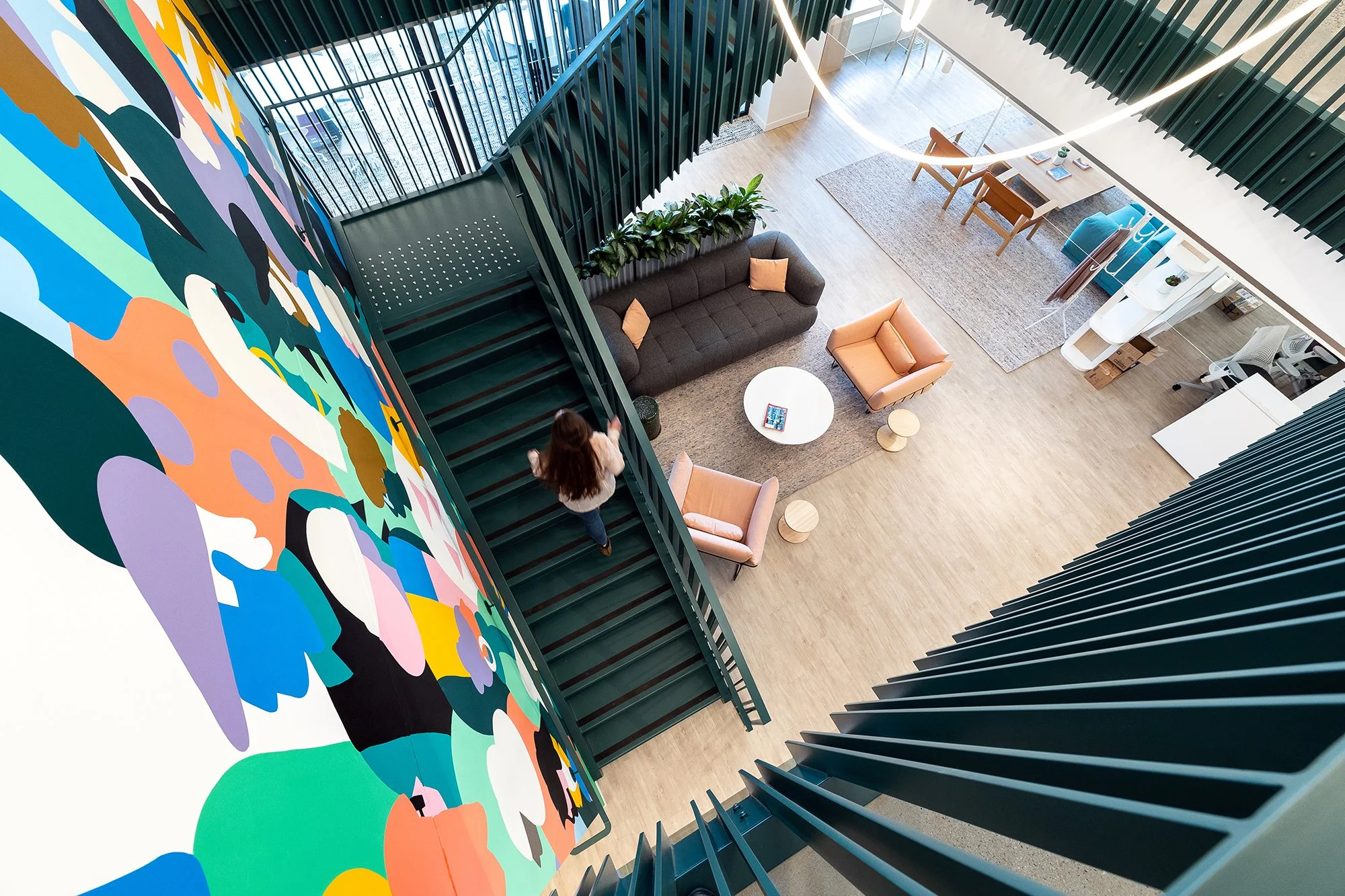 Overhead view of a modern, brightly lit living space with a staircase, colorful abstract wall art, seating area with two armchairs, sofa, and a small round table. A person with long brown hair is walking up the stairs.