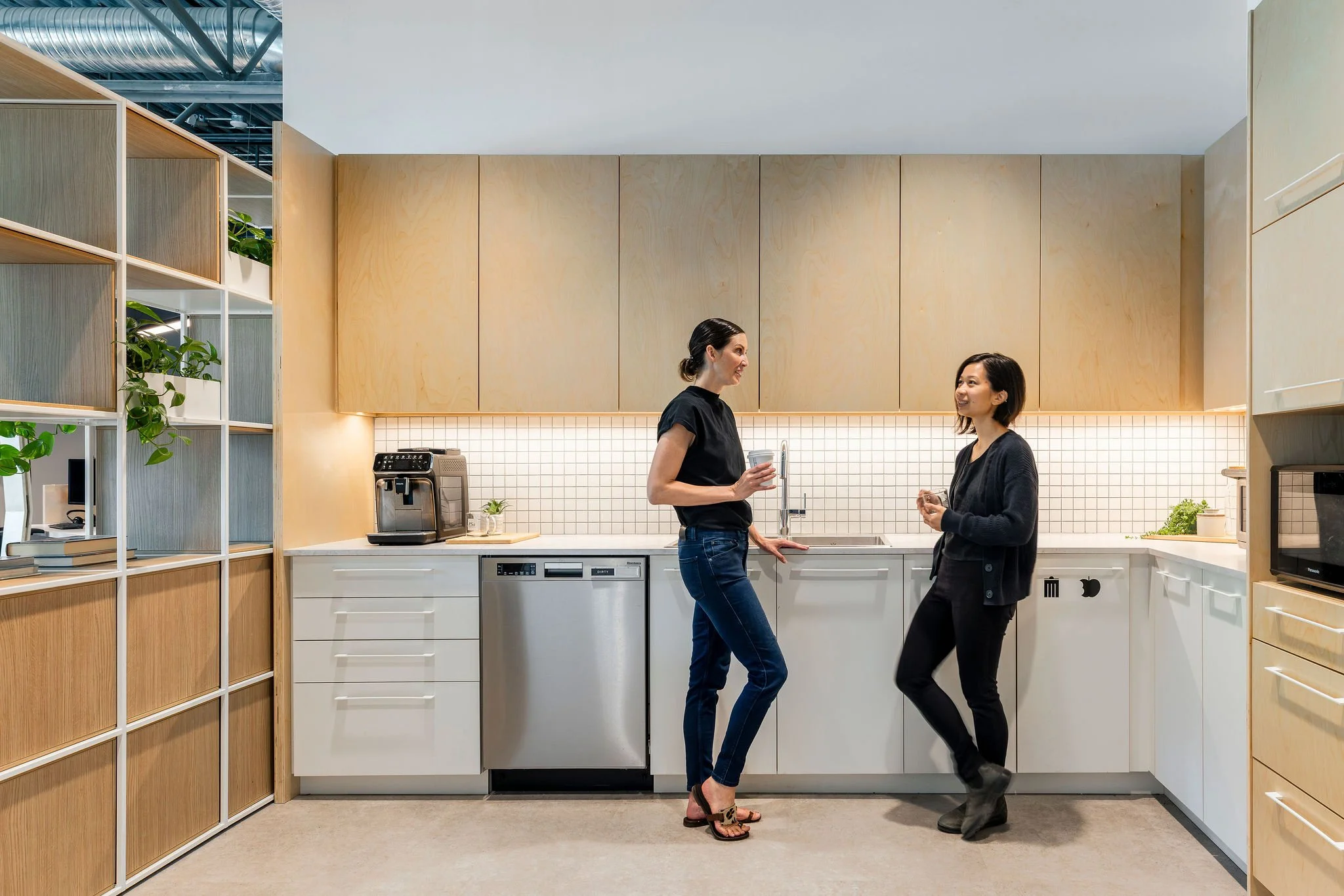 Two women are having a conversation in a modern kitchen. One woman is holding a cup, and the other is smiling and looking at her. The kitchen has wooden cabinets, a white countertop, and some plants on the shelves.