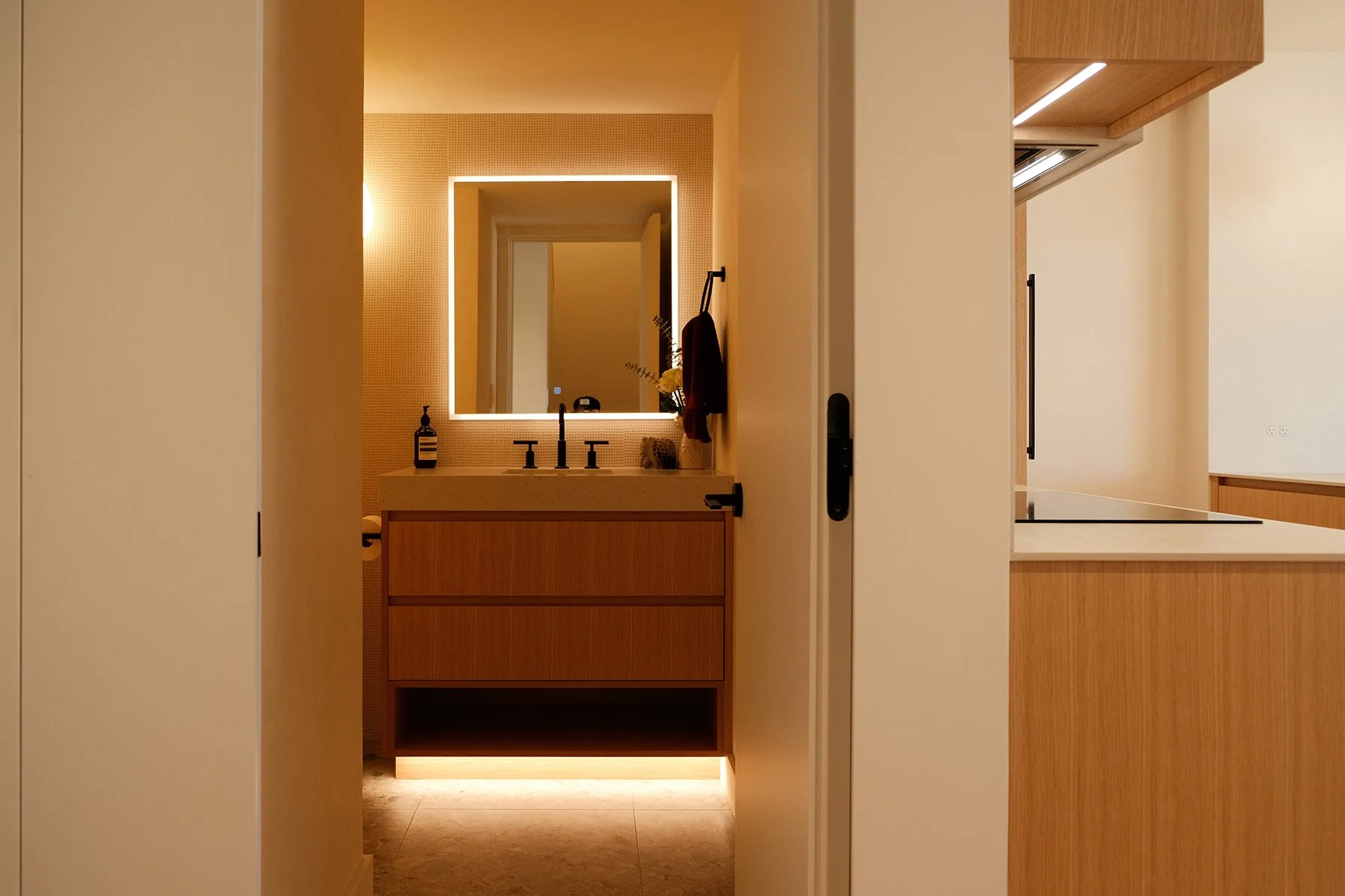 Modern bathroom with a lit mirror, a wooden vanity, a black faucet, and bottles on the sink, visible through an open door from a kitchen area with wooden cabinets.