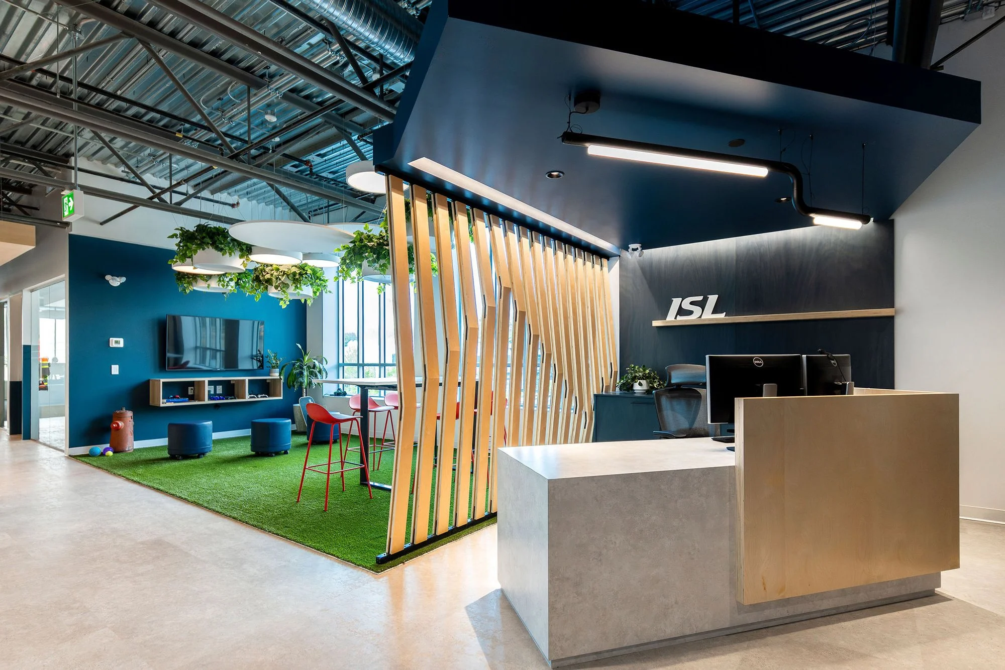Modern office interior with a reception desk, wooden room dividers, a sitting area with a green carpet, chairs, and a television