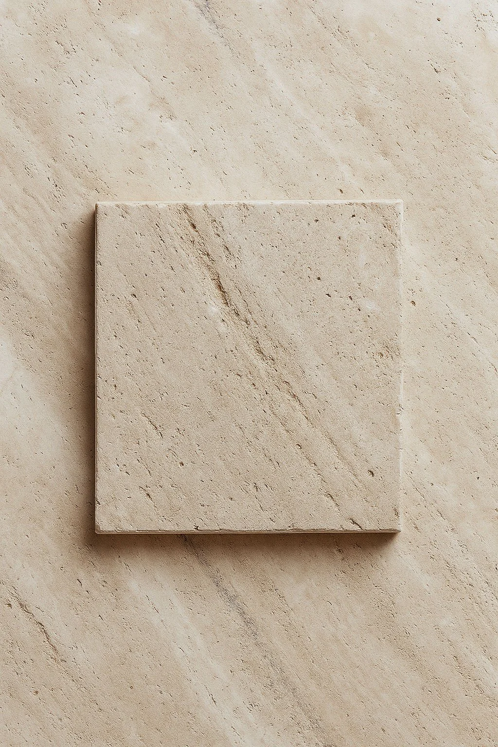 A square beige marble tile with natural veining, placed on a matching beige marble surface.