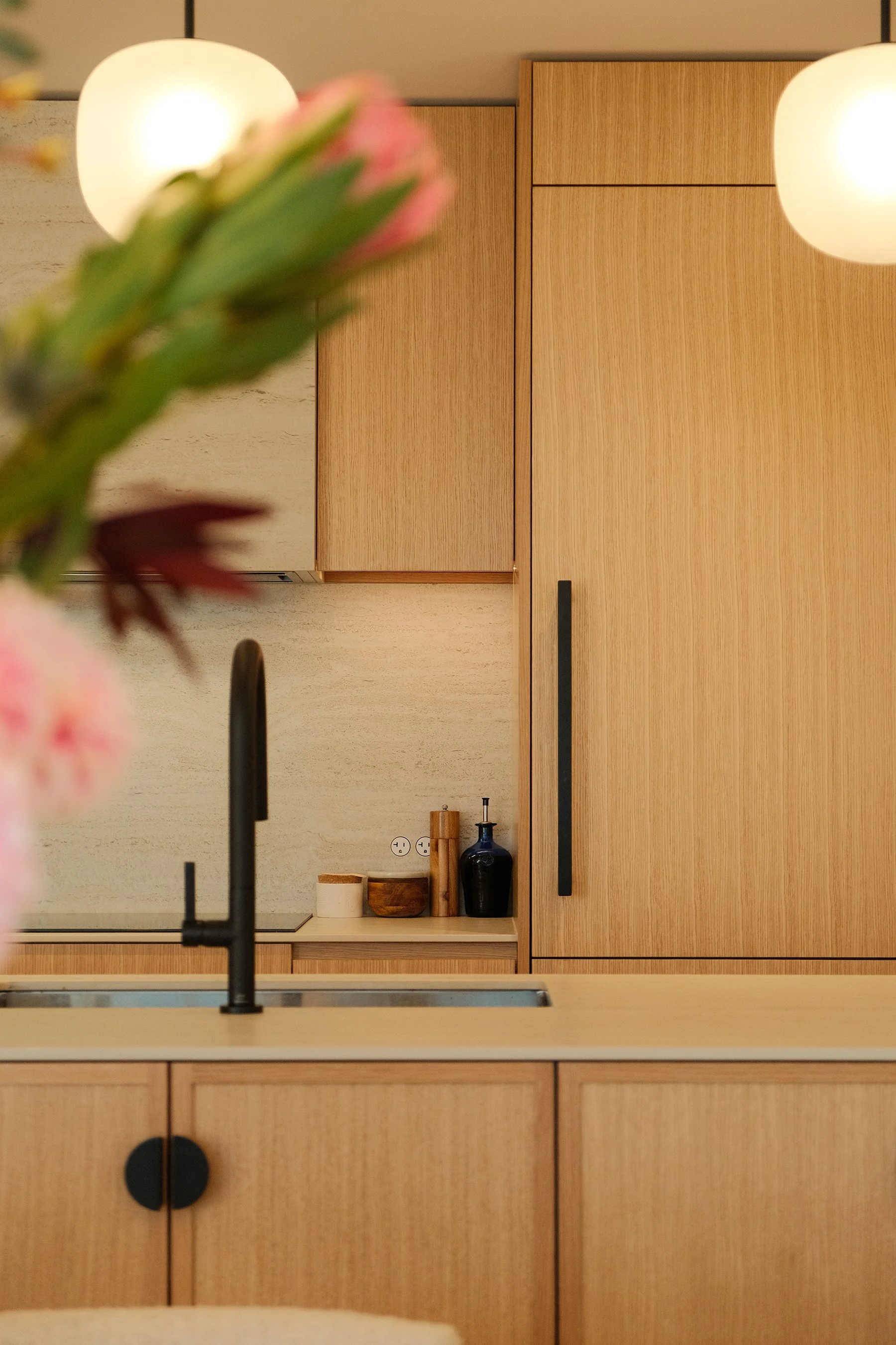 A modern kitchen with light wood cabinets, a black faucet, and minimal decor, with a flower in the foreground.