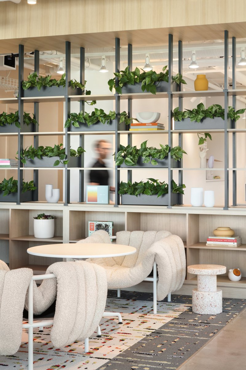 A modern interior with a beige armchair and sofa, a round white table, decorative books, and potted plants arranged on a black and beige shelving unit dividing the space. There are decorative vases and a small round table in the background.