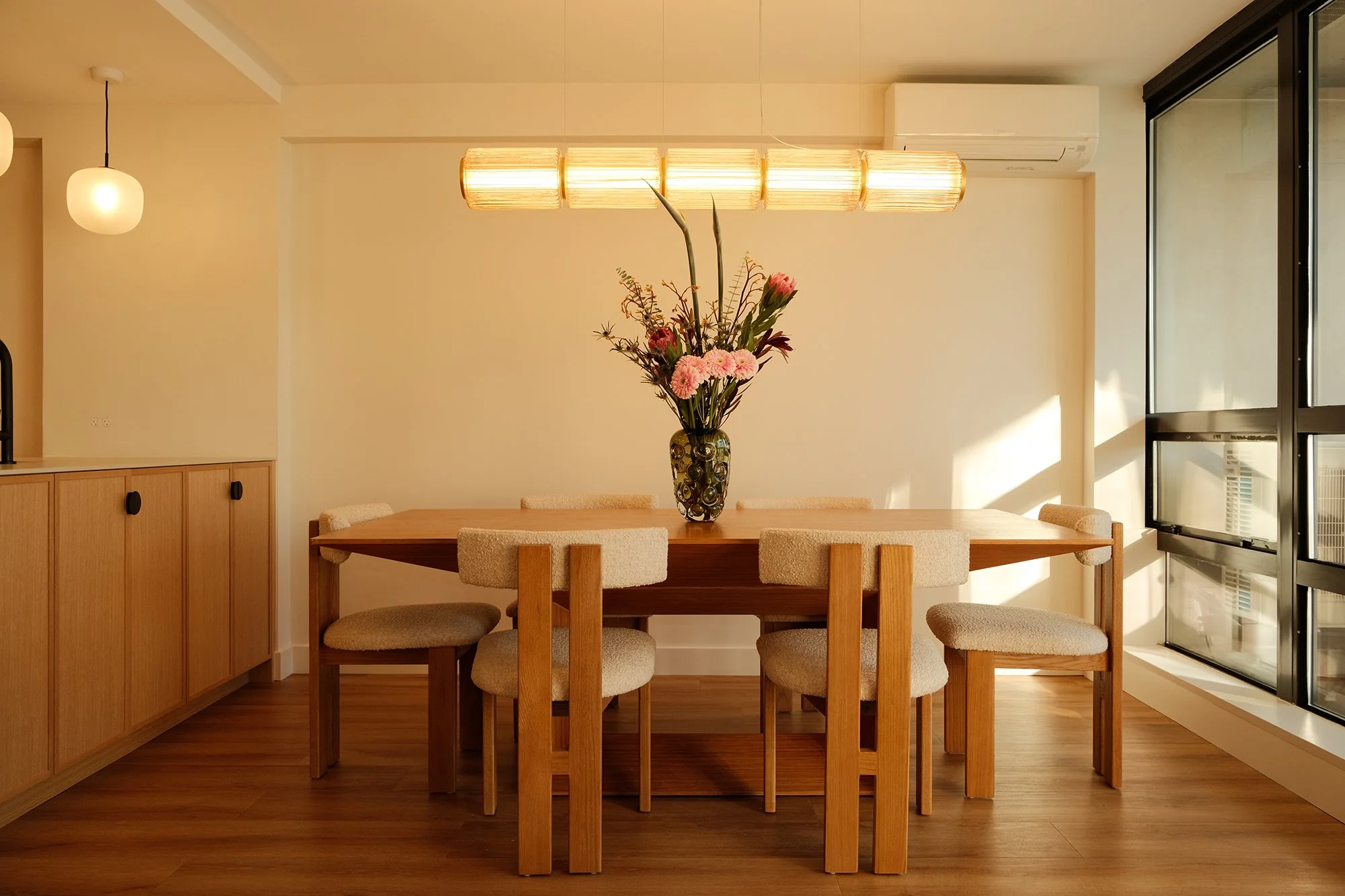A dining room with a wooden table and beige upholstered chairs, a floral arrangement in a dark vase on the table, a modern hanging light fixture, and an air conditioning unit on the wall.