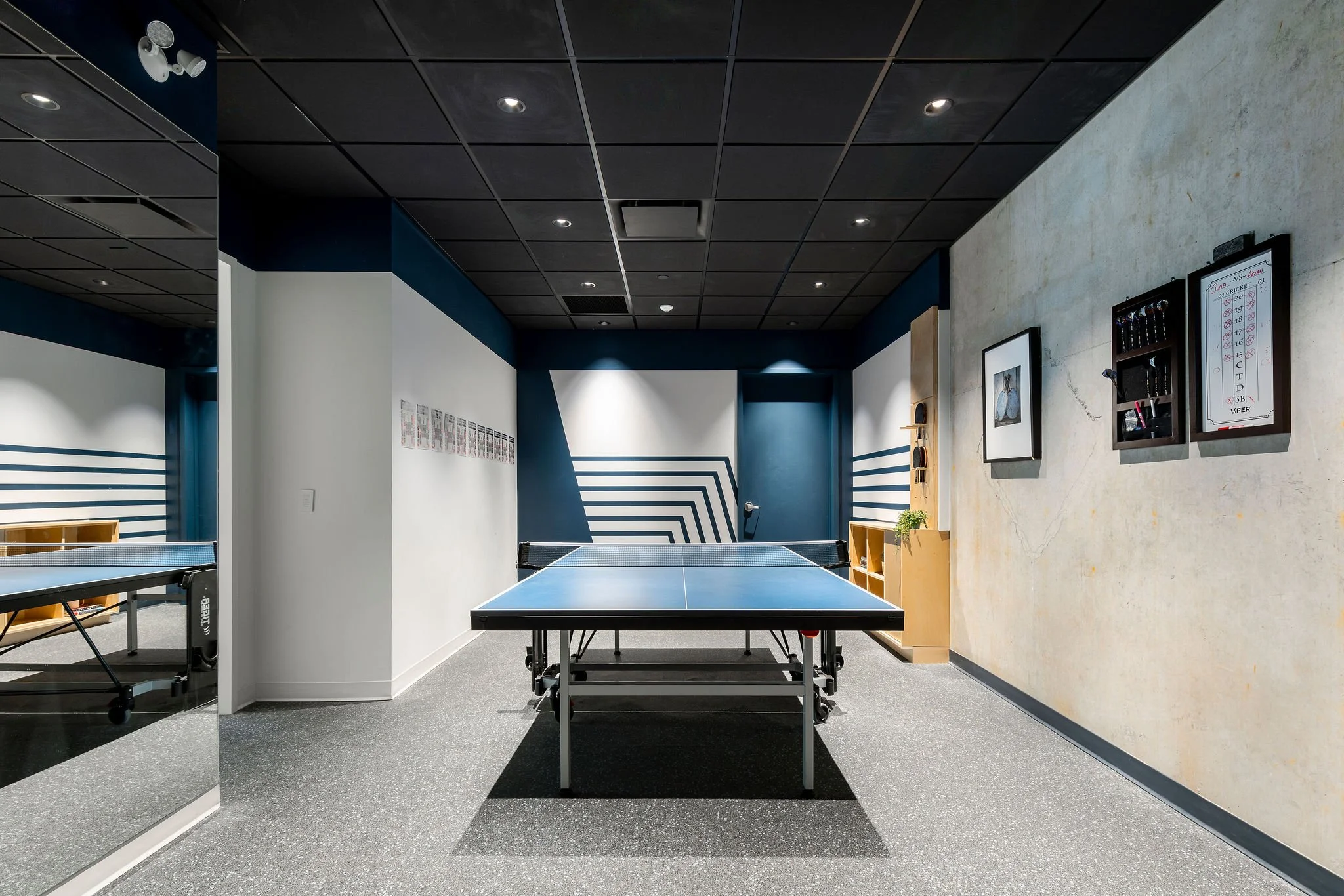 Indoor recreational space with a ping pong table at the center, blue and white walls, and a black ceiling with ceiling lights.