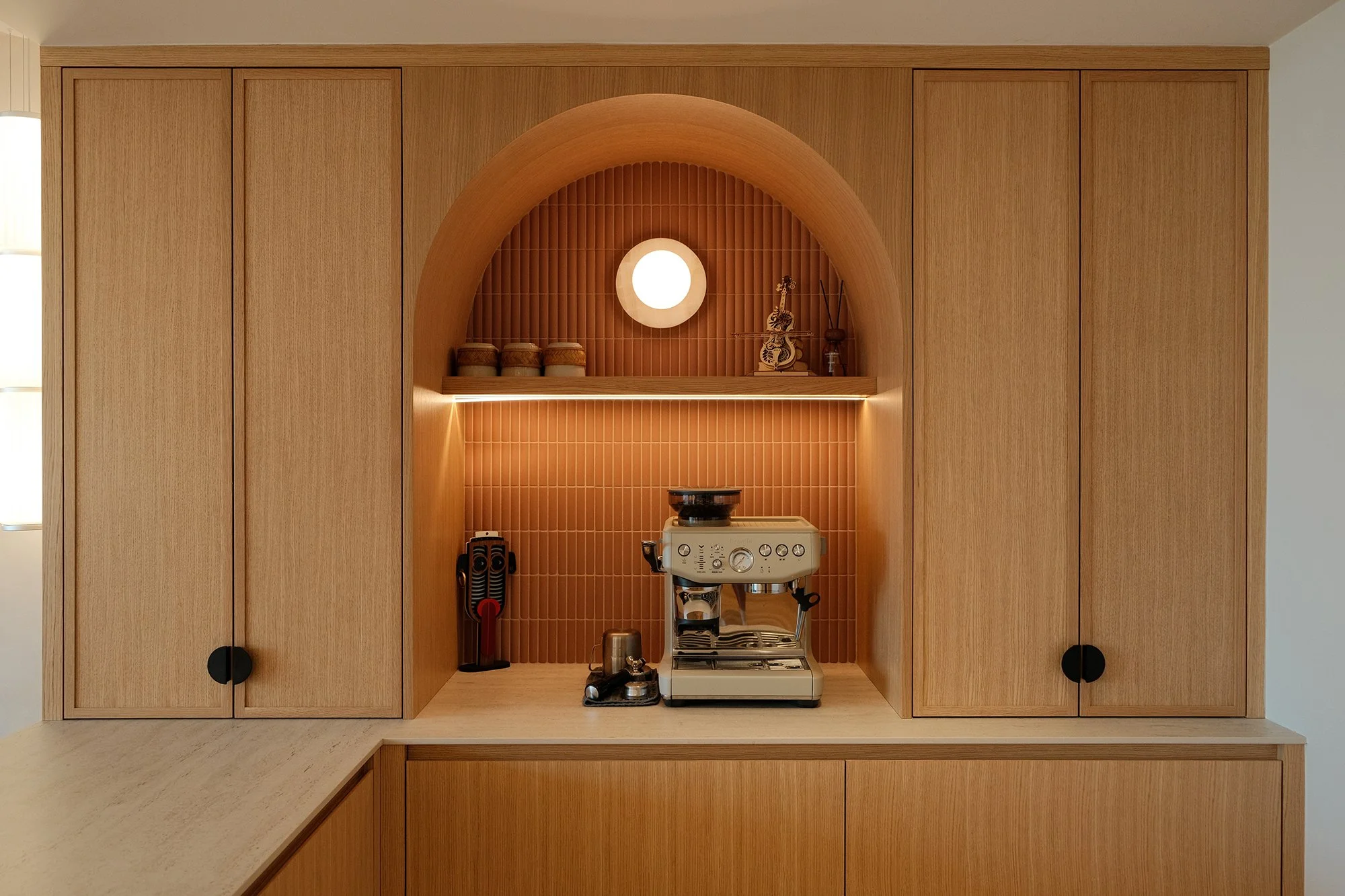 Built-in kitchen cabinet with a coffee machine, a scale, and decorative items on a shelf, illuminated by warm lighting.