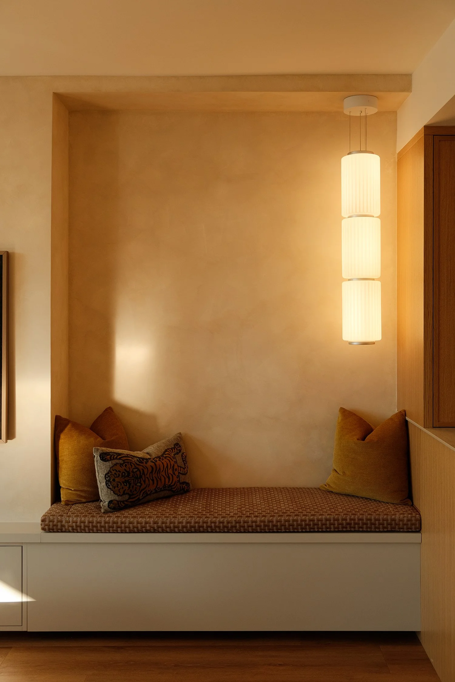 A cozy seating nook with cushions and pillows, illuminated by a hanging cylindrical light fixture, against a beige wall with wooden accents.