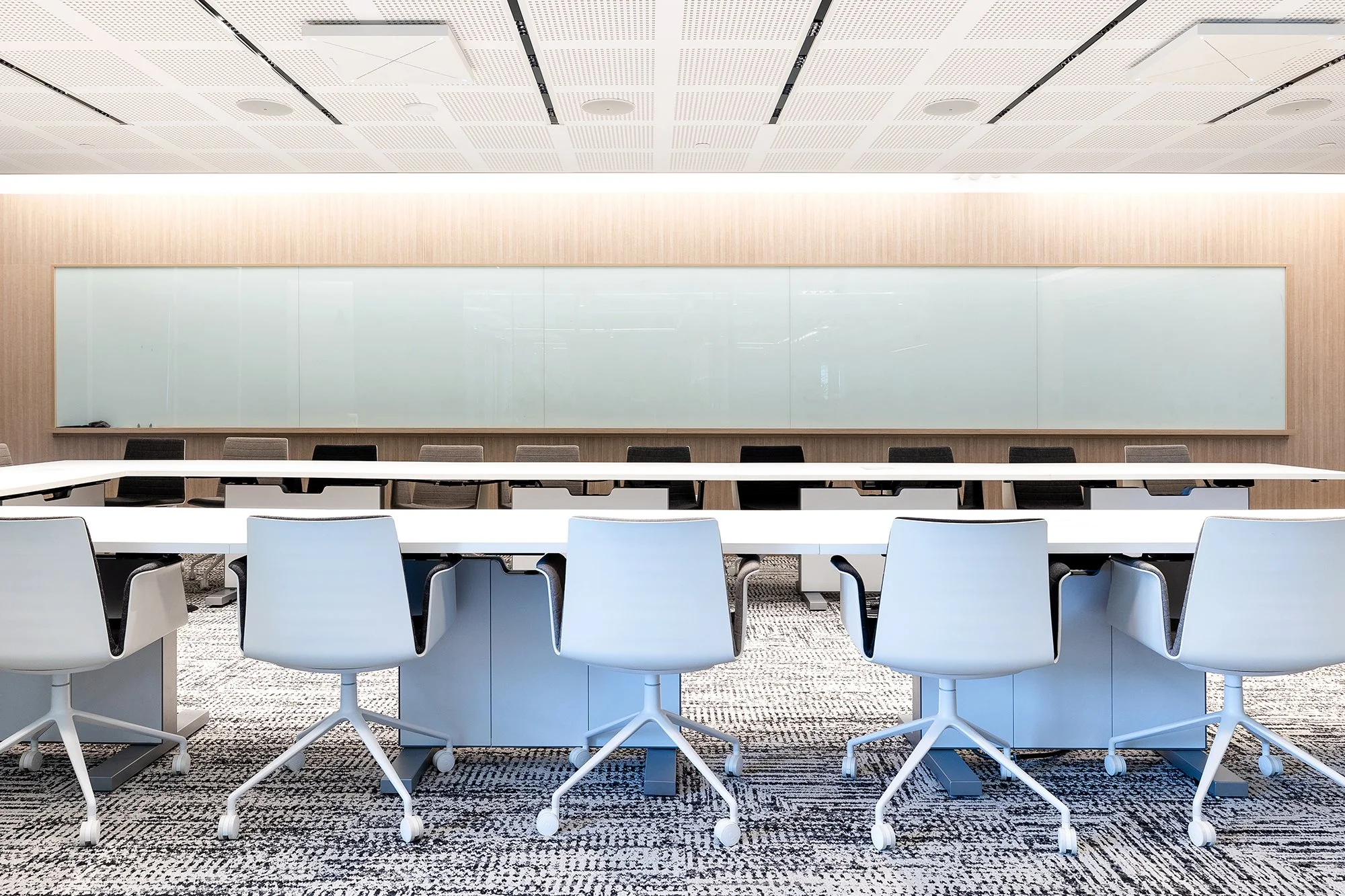 A modern conference room with white chairs around a long white table, facing a large whiteboard on a light wood wall. The ceiling has white panels with holes and black strips, and the floor is carpeted in a black and white pattern.