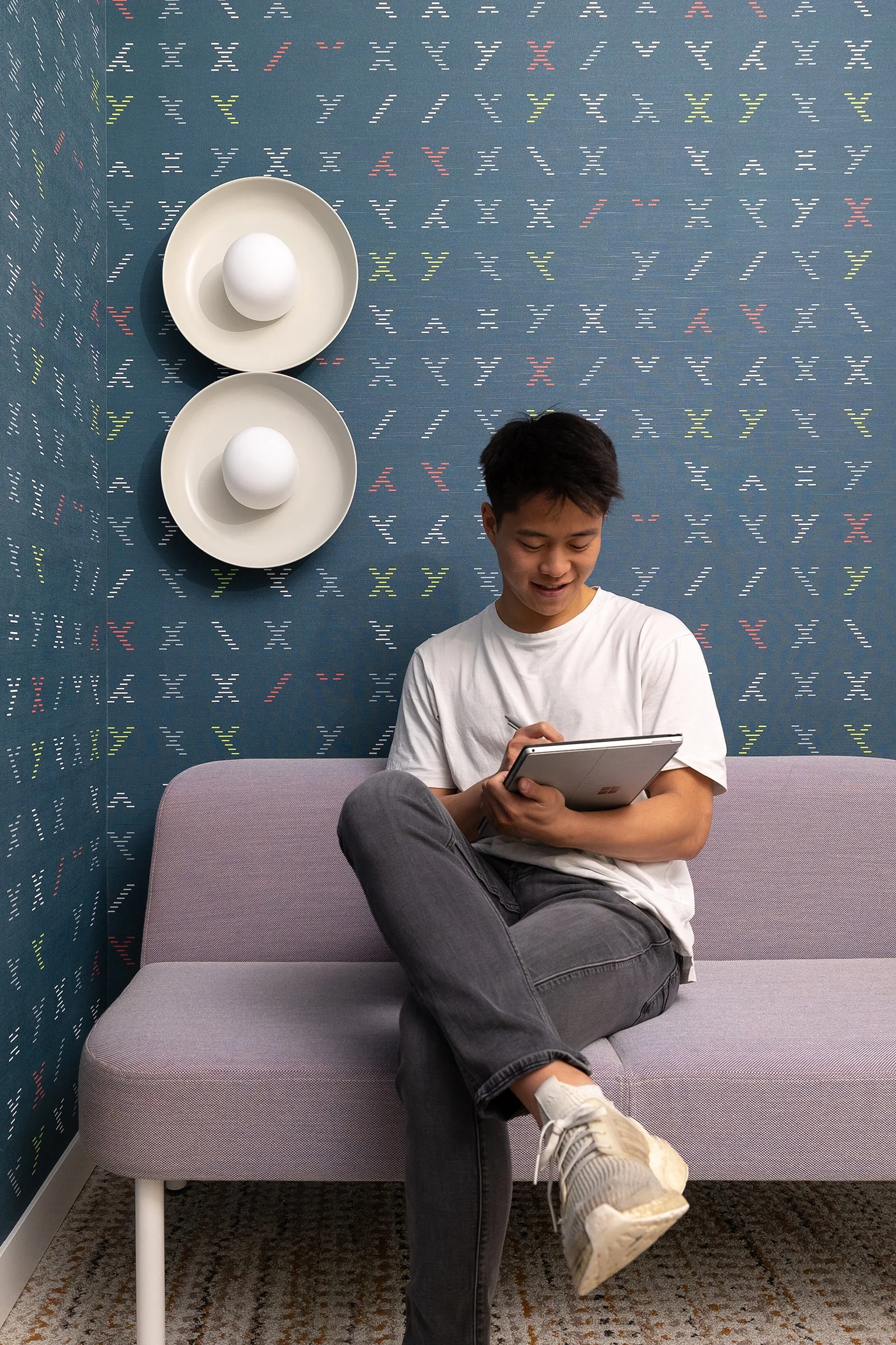 A young man sitting on a gray sofa with his legs crossed, looking at a tablet device while smiling. He is wearing a white t-shirt, dark jeans, and sneakers. The background features a blue wall with colorful, dotted 'X' and 'Y' patterns, and two white plates each with a white egg on top are mounted on the wall behind him.