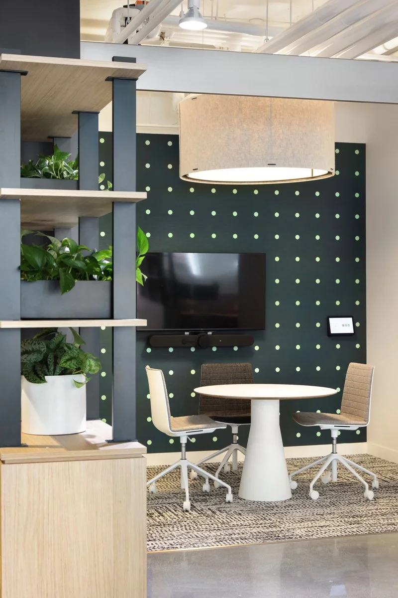 Modern office meeting area with a round white table, three office chairs, a wall-mounted TV, and a large circular ceiling light.