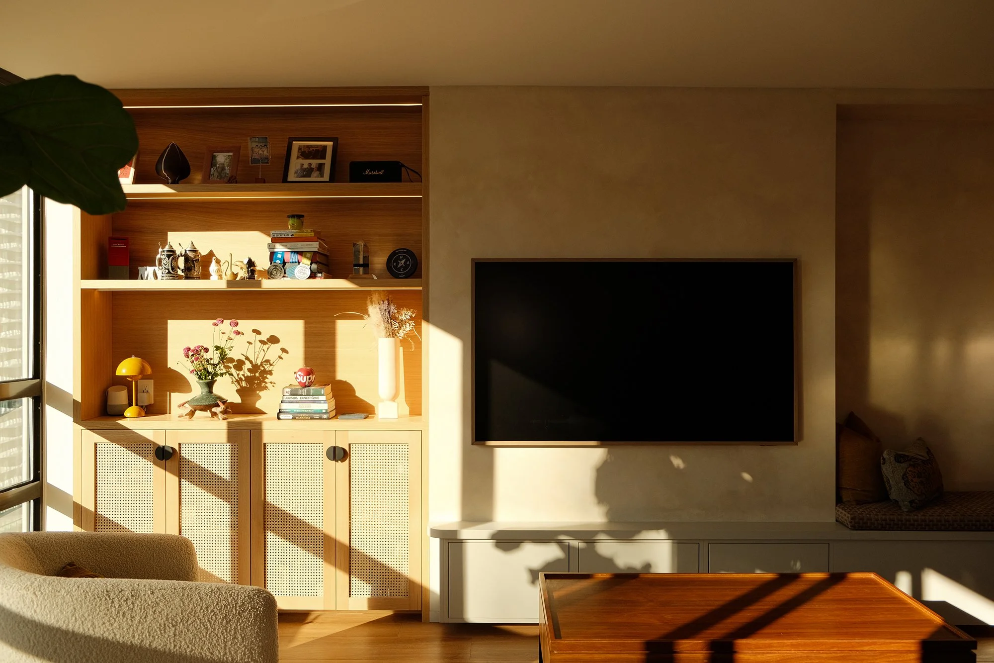 Living room with wooden shelving, decorative items, books, and a large flat-screen TV, illuminated by sunlight.