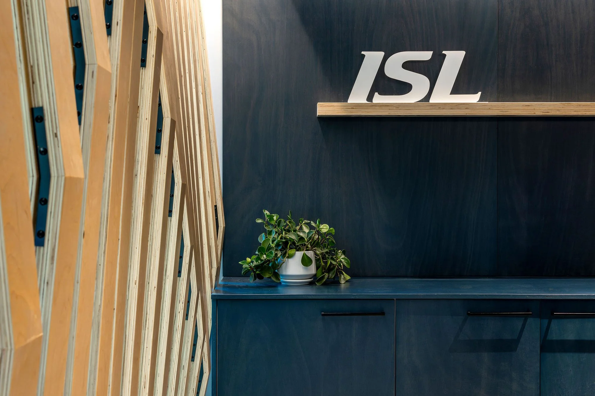 Interior space with wooden lattice divider on the left, dark gray wall on the right with white 3D letters spelling 'ISL,' a small dark cabinet, and a green leafy potted plant on a gray surface.