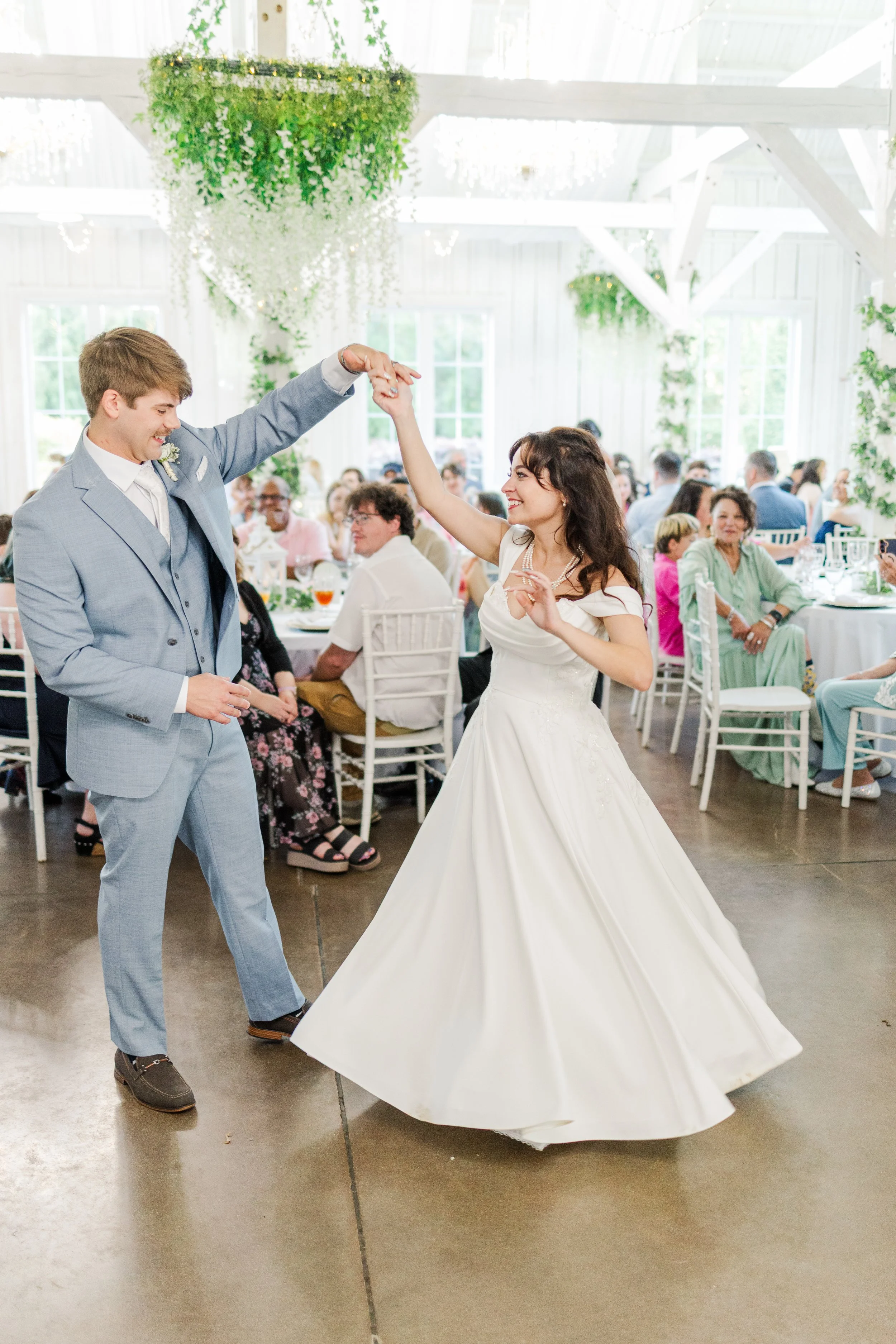 A bride and groom dancing at their wedding reception inside a bright, decorated venue. The groom is wearing a light gray suit, and the bride is in a white wedding gown with a full skirt. There are guests seated at tables in the background, and the ve
