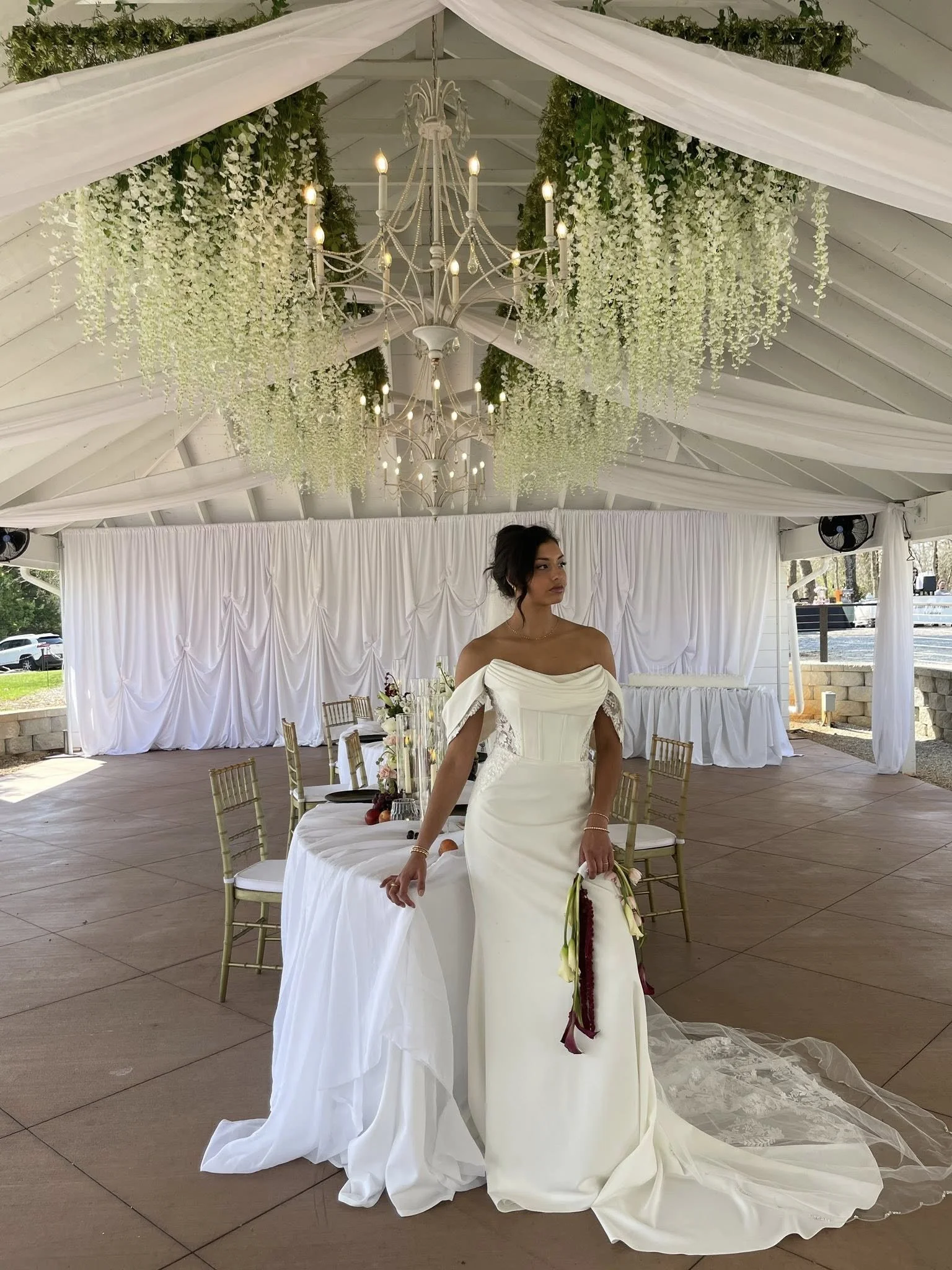 A woman in a white wedding dress is standing under a decorated canopy with white drapery, hanging white flowers, and chandeliers. There are tables with chairs around her set for a wedding reception.