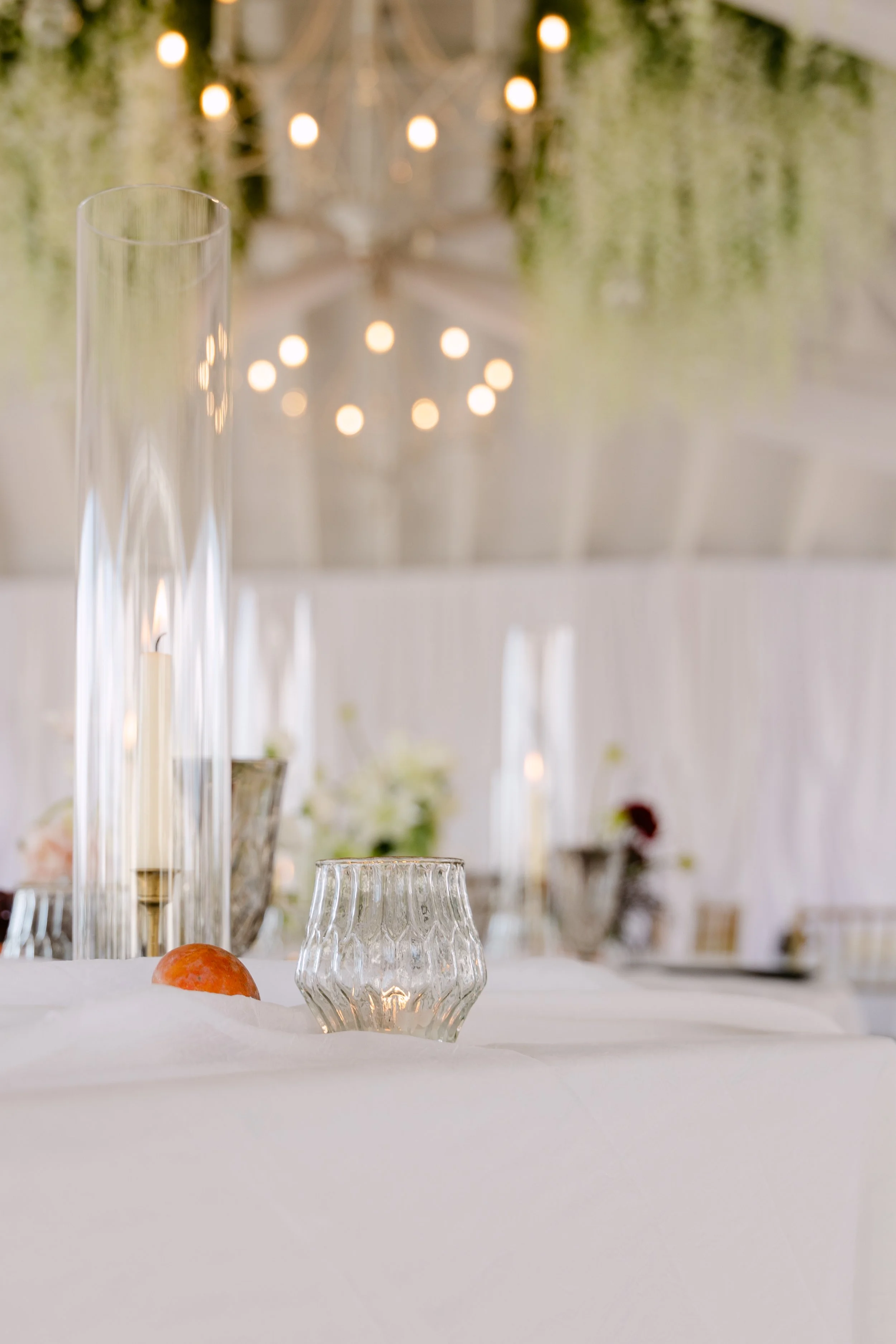 Glass candle holders and a small orange decorative item on a table, with blurred elegant lighting and greenery in the background.