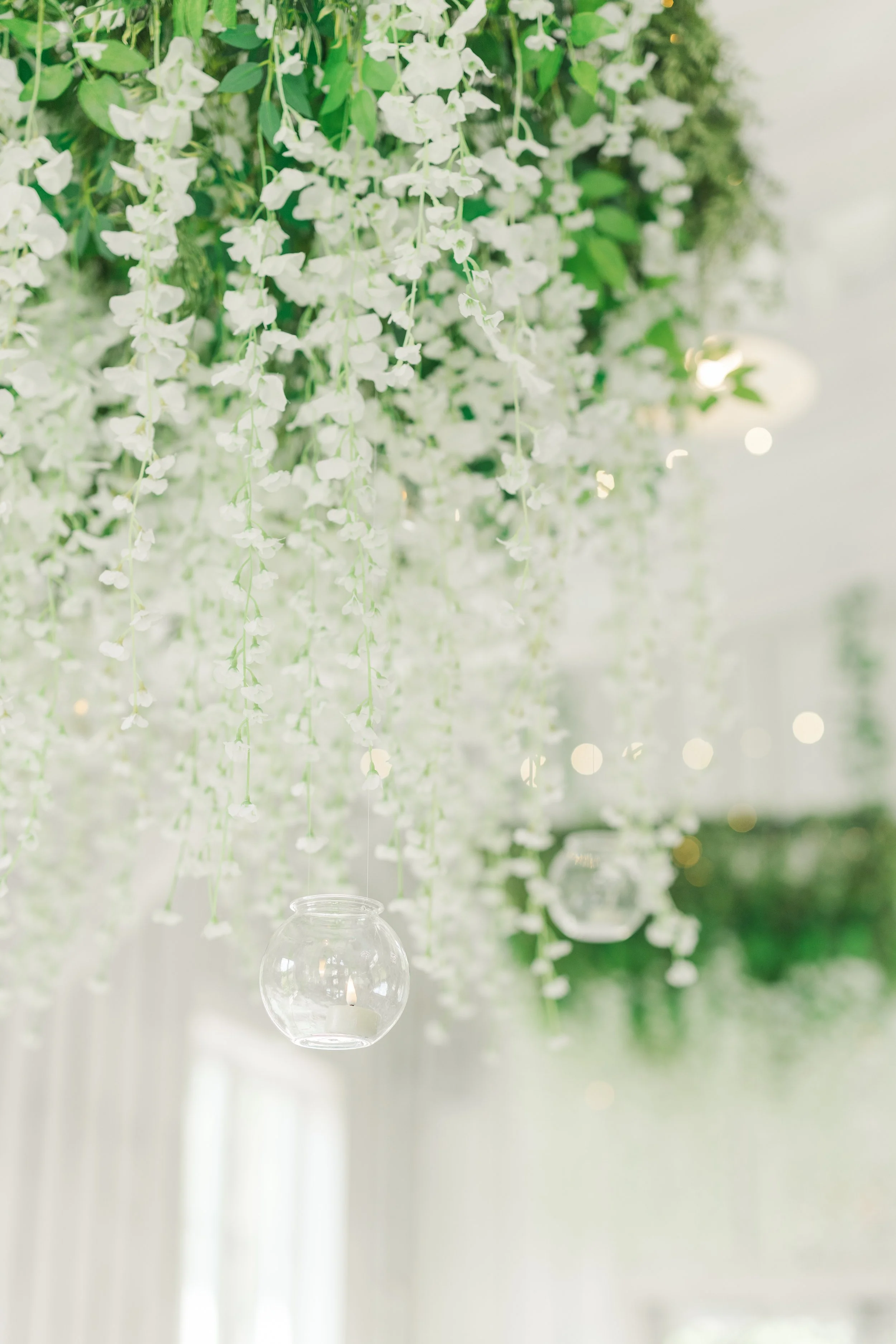 Hanging white flowers and glass candle holders with lit candles in a softly lit space.