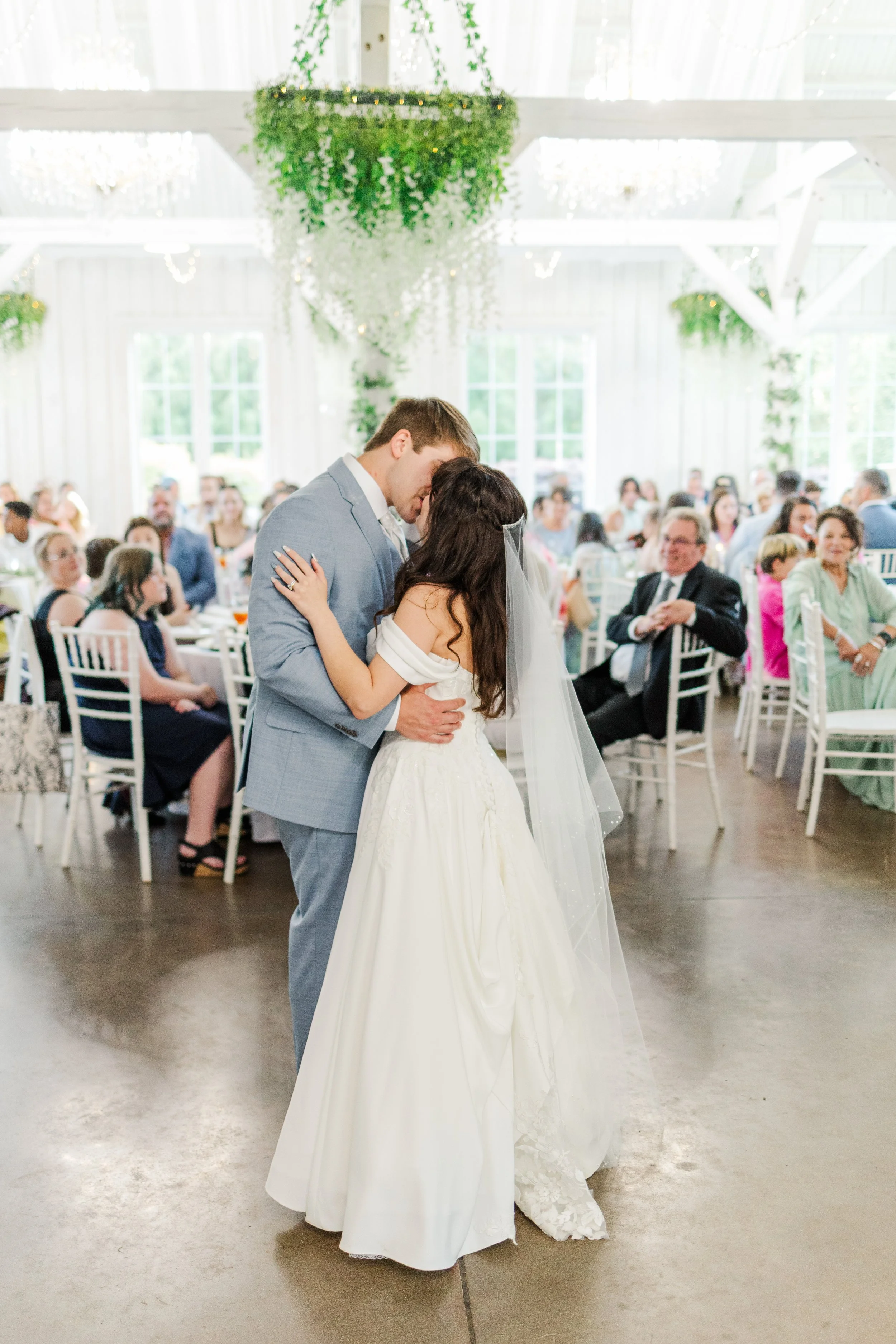 A bride and groom sharing a dance during their wedding reception in a bright, decorated hall, with guests seated at tables in the background.
