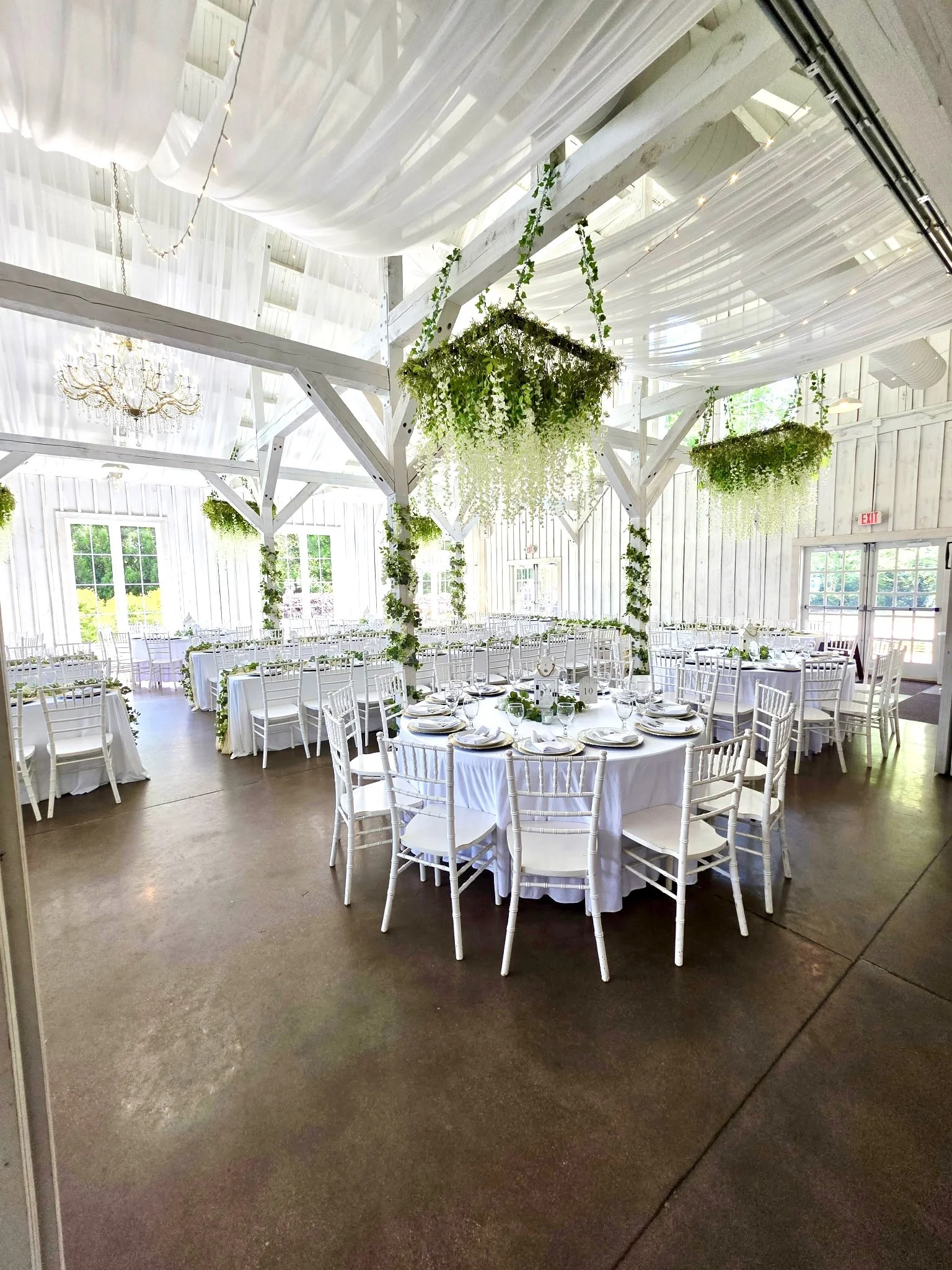 Elegant reception hall decorated with white drapery and hanging greenery, round tables with white tablecloths, white chairs, and natural lighting through windows.