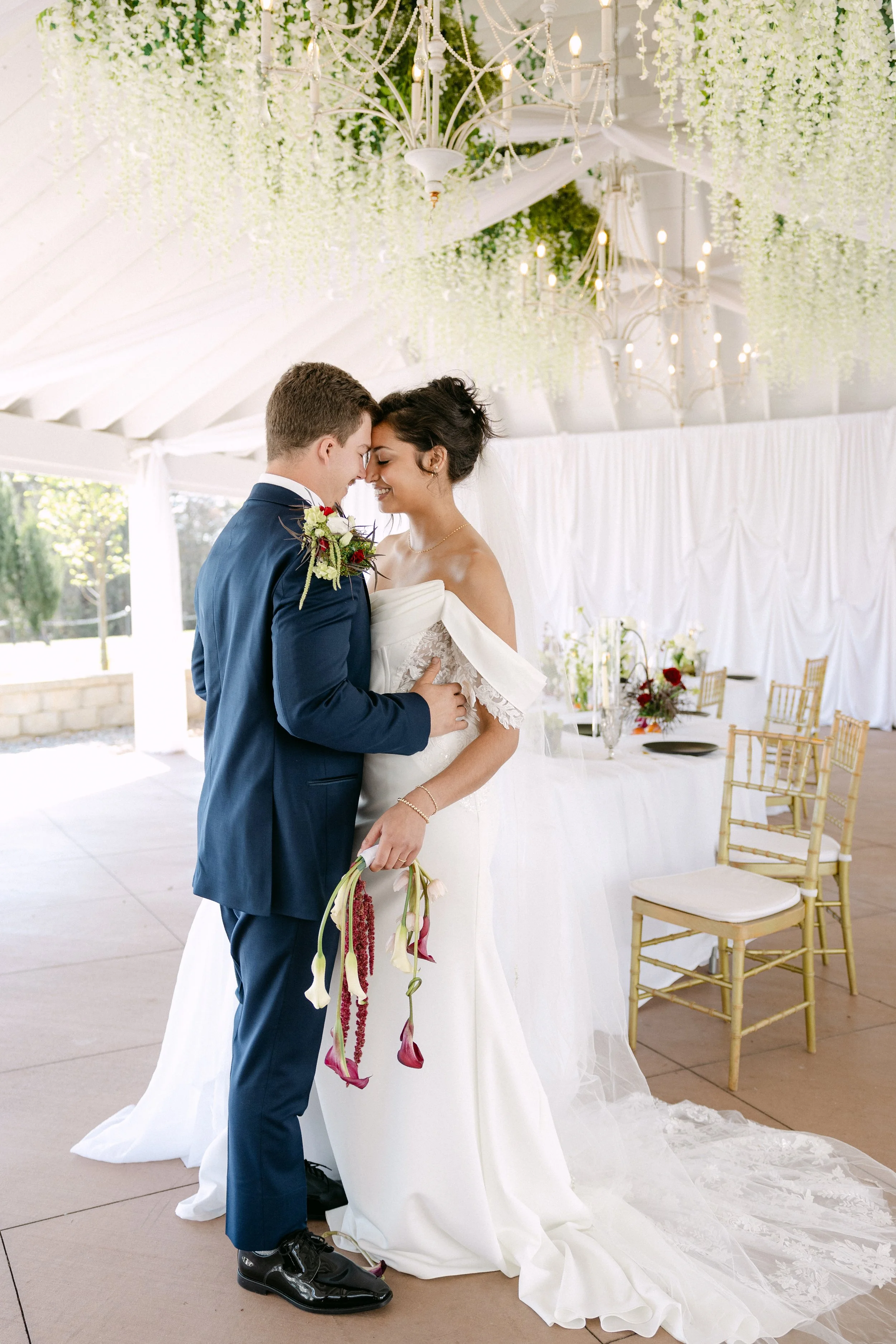 A bride and groom standing close together, touching foreheads and smiling during a wedding ceremony inside a decorated venue with white drapes, chandeliers, and chairs.