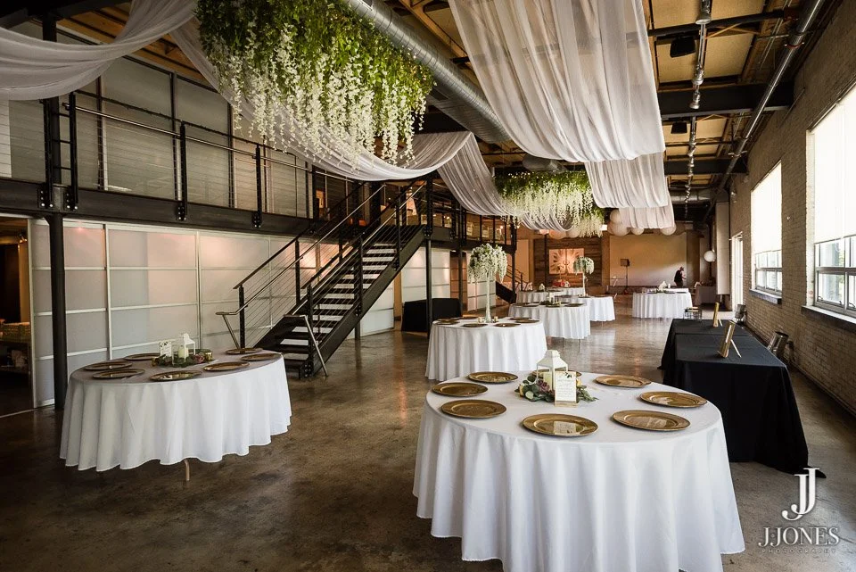 An event space decorated with white drapery and greenery, with several round tables covered in white tablecloths and gold plates, set for a formal event or wedding reception. The space includes a staircase, large windows, and a brick wall.