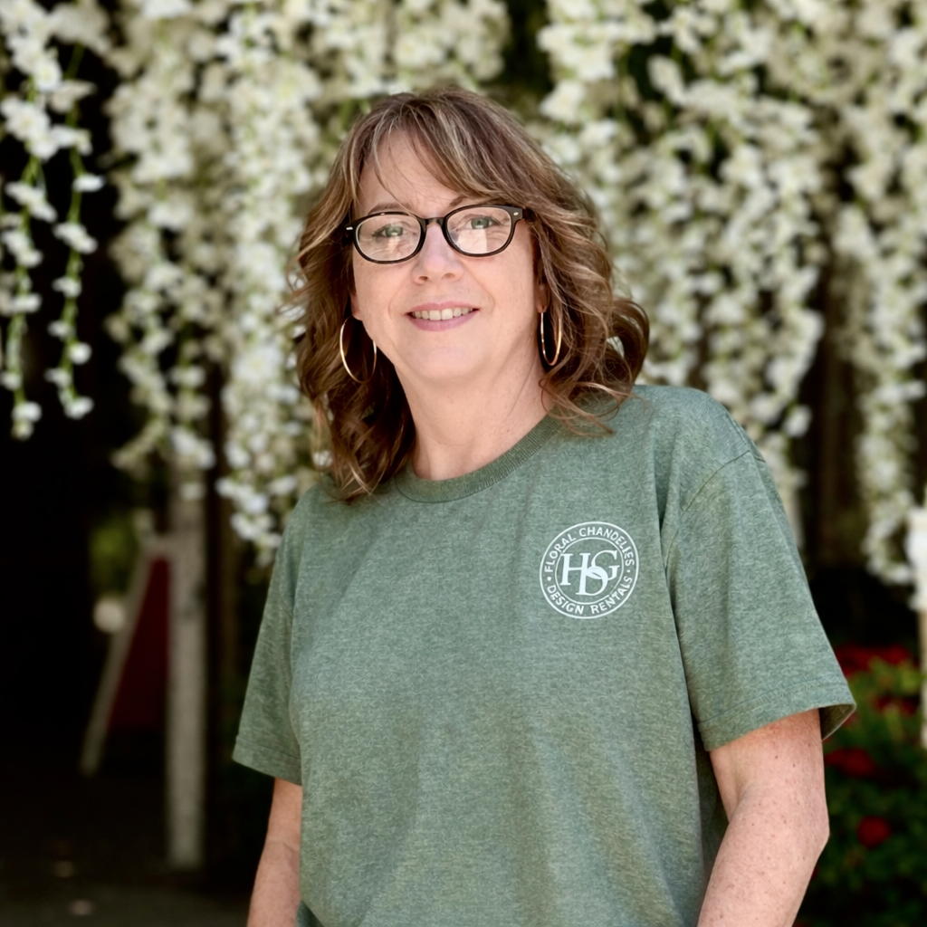 A woman with glasses and curly hair, wearing a green t-shirt with a logo, standing outdoors with blooming white flowers in the background.