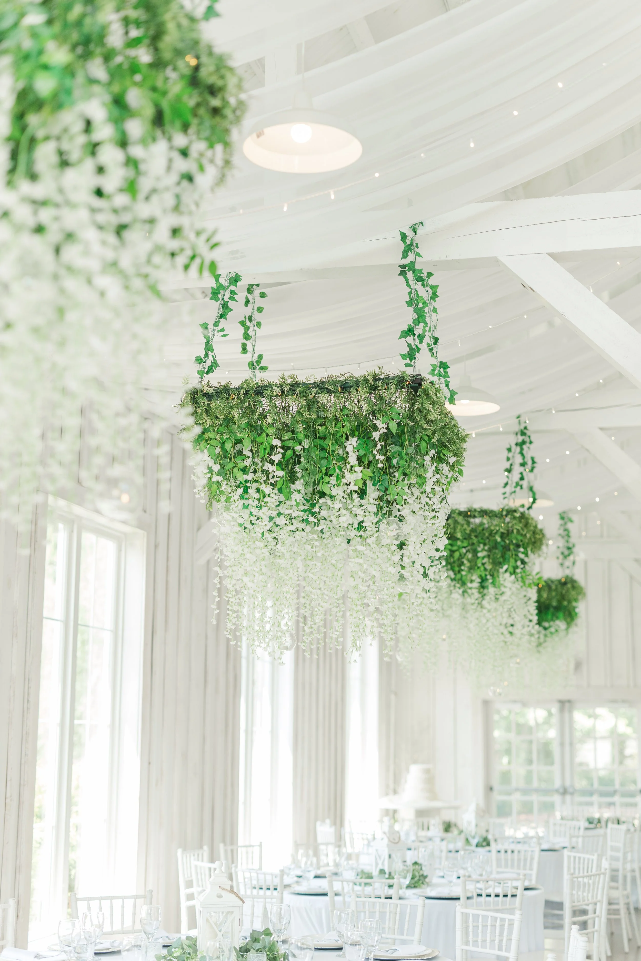 Indoor wedding reception with white chairs, tables, and hanging greenery installation, bright natural light, white wooden walls and ceiling.