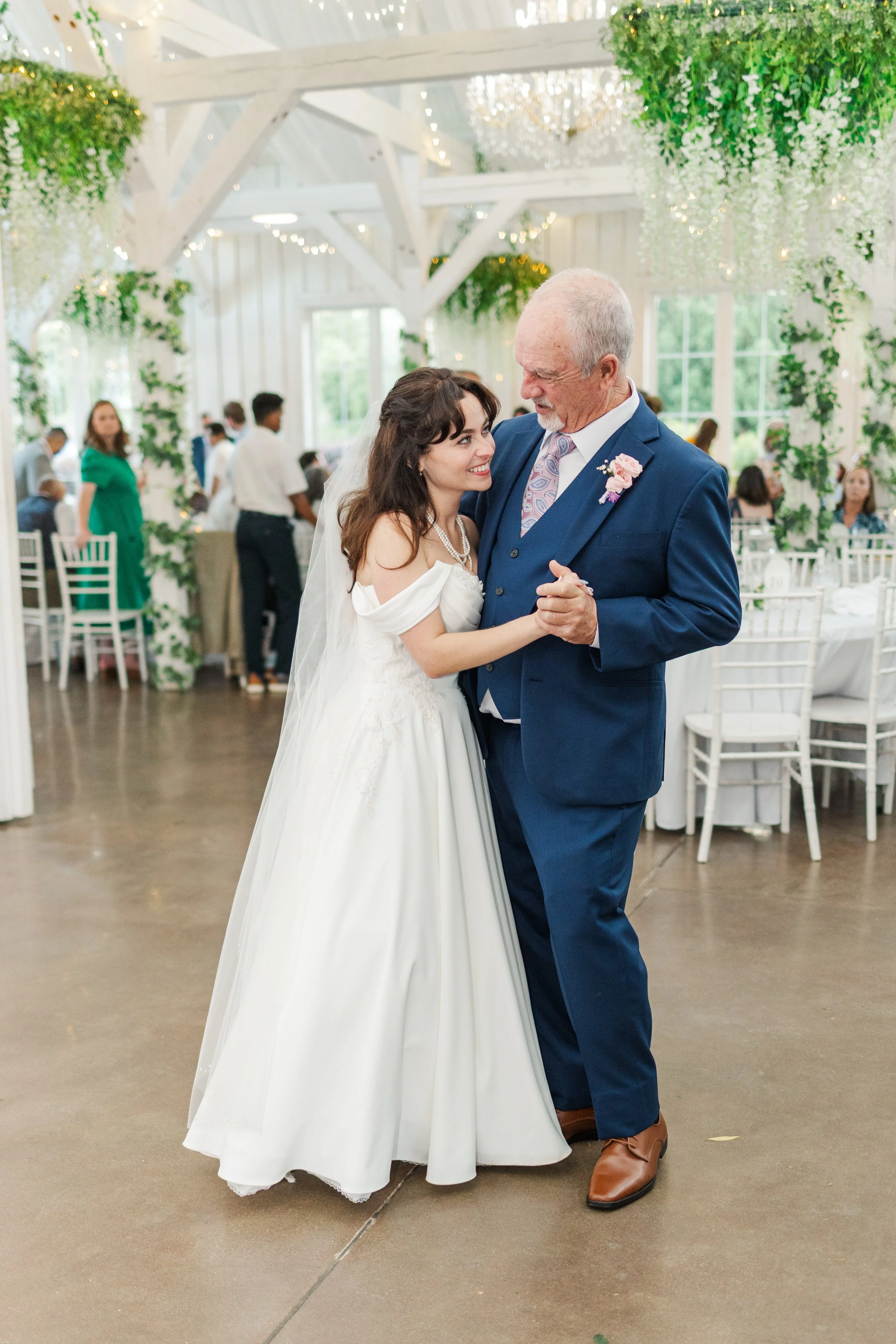 A bride wearing a white off-the-shoulder wedding dress with a pearl necklace dances with an elderly man in a blue suit at a wedding reception with white chairs, greenery, and string lights.