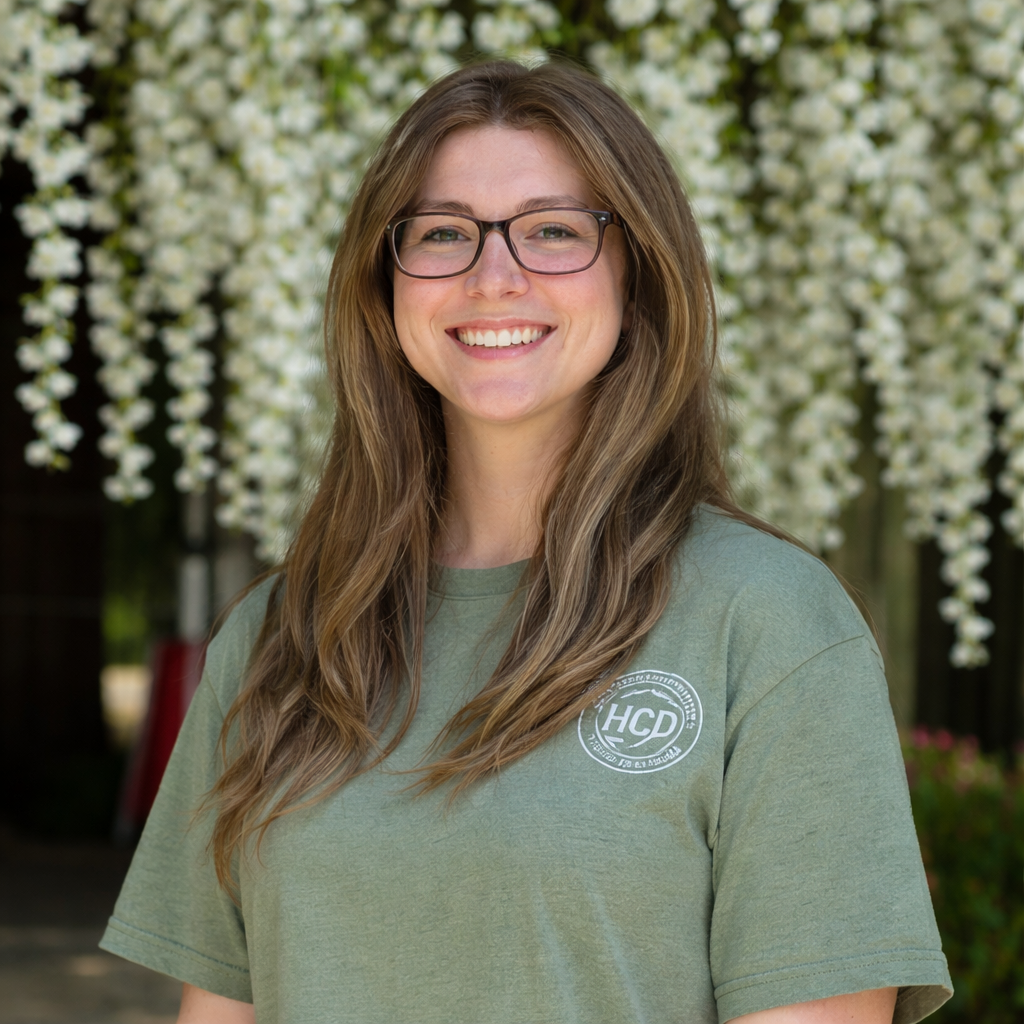 A young woman with glasses and long brown hair smiling outdoors in front of a backdrop of white flowering trees. She is wearing an olive-green T-shirt with a circular logo on the chest.